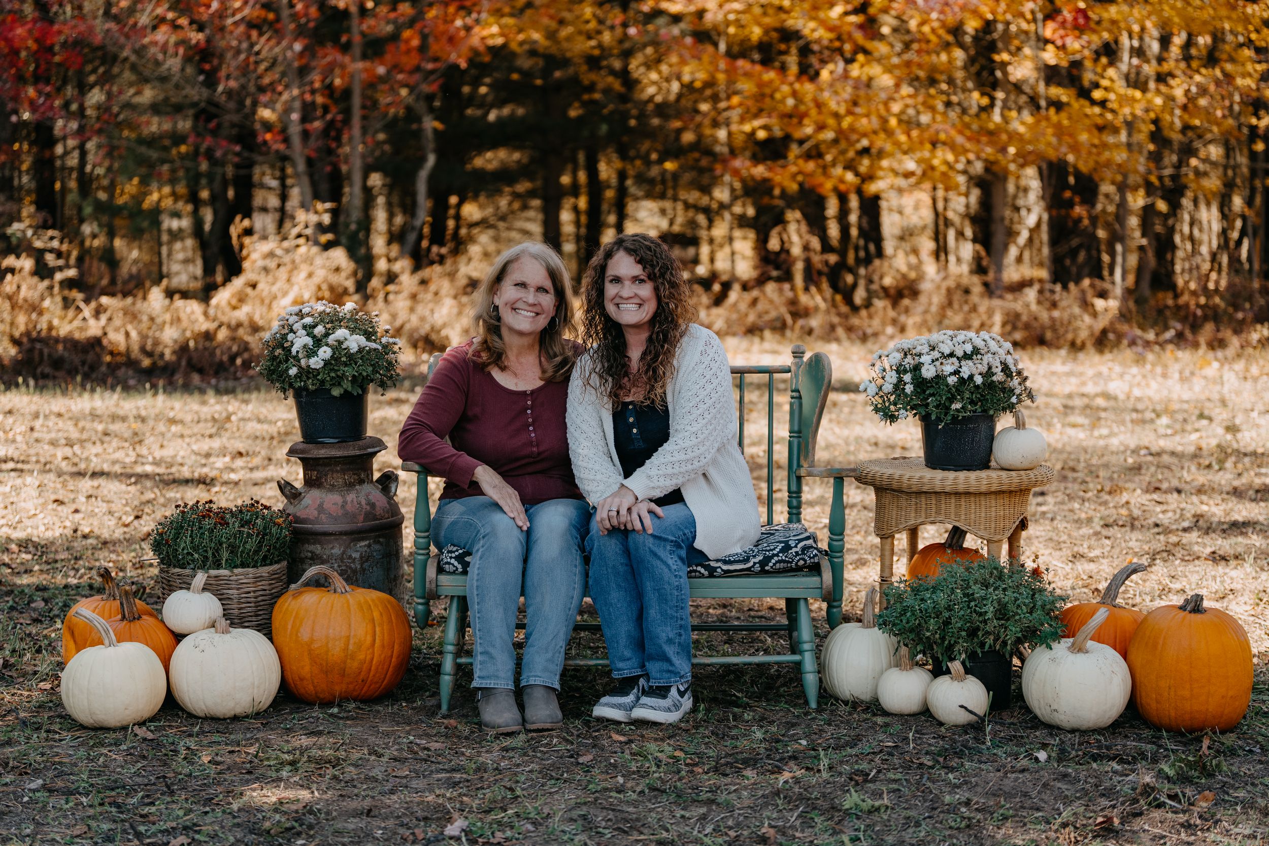 Two people sit on a bench surrounded by orange and white pumpkins and potted plants in an autumn outdoor setting.
