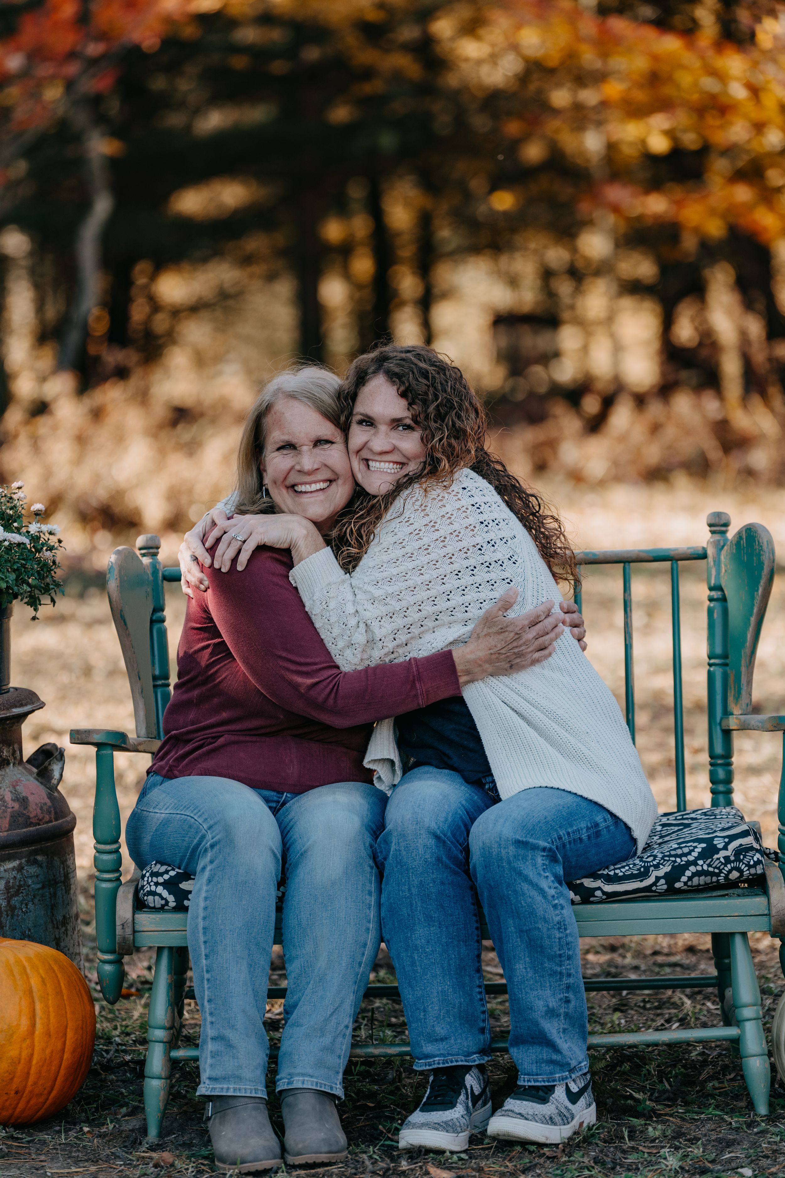 Two people share a warm embrace while sitting on blue chairs outdoors with autumn foliage and a pumpkin in the background.