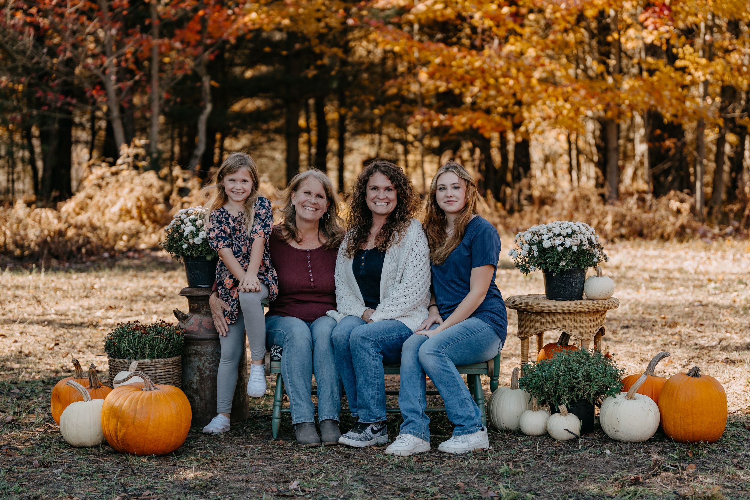 A group sits on a bench surrounded by autumn pumpkins and fall foliage in a rustic outdoor setting.