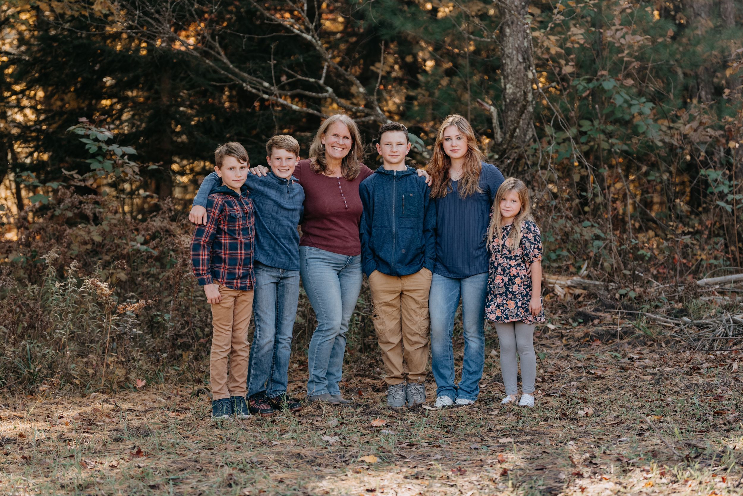 A group of people pose together in a casual outdoor autumn setting among fallen leaves and trees.