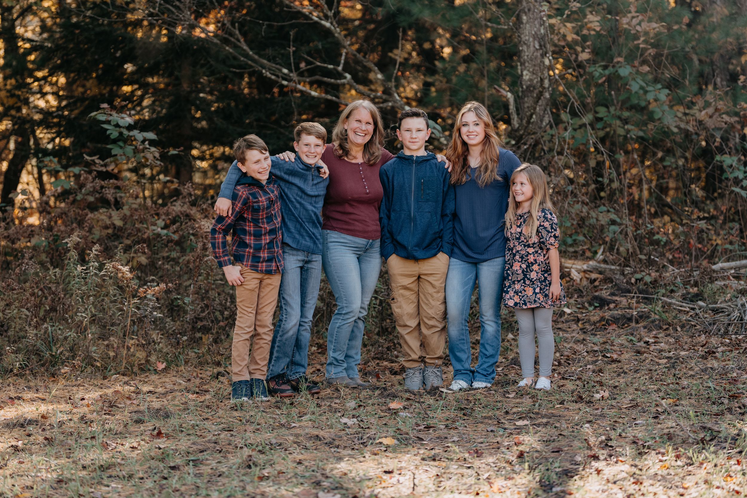 Outdoor autumn family portrait with people wearing coordinated casual outfits against a wooded background.