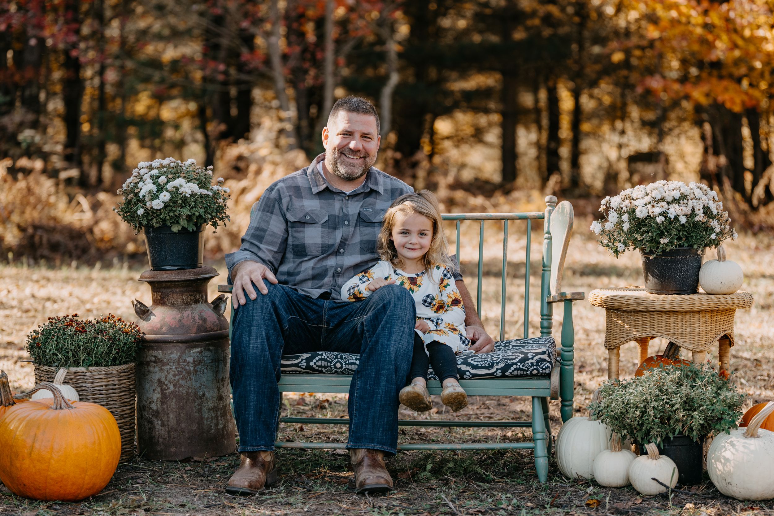 A rustic autumn outdoor photo session with people sitting on wooden benches among orange foliage and pumpkin decorations.