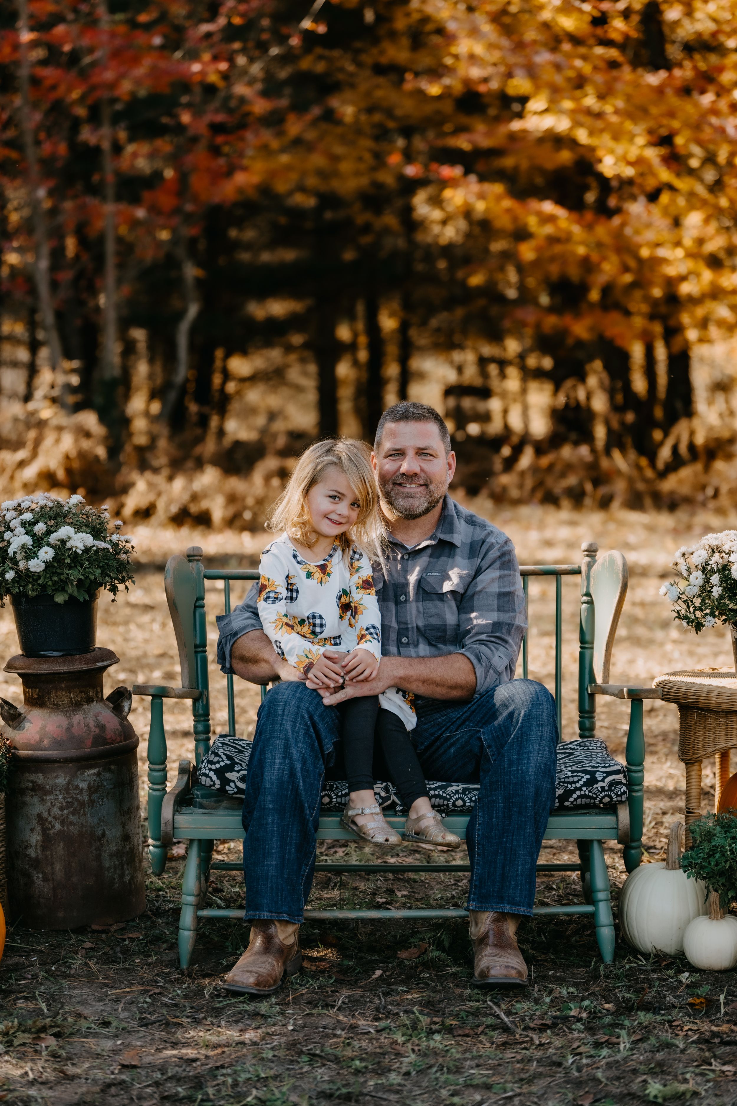 A rustic autumn scene with a bench decorated with pumpkins and flowers in front of colorful fall foliage.
