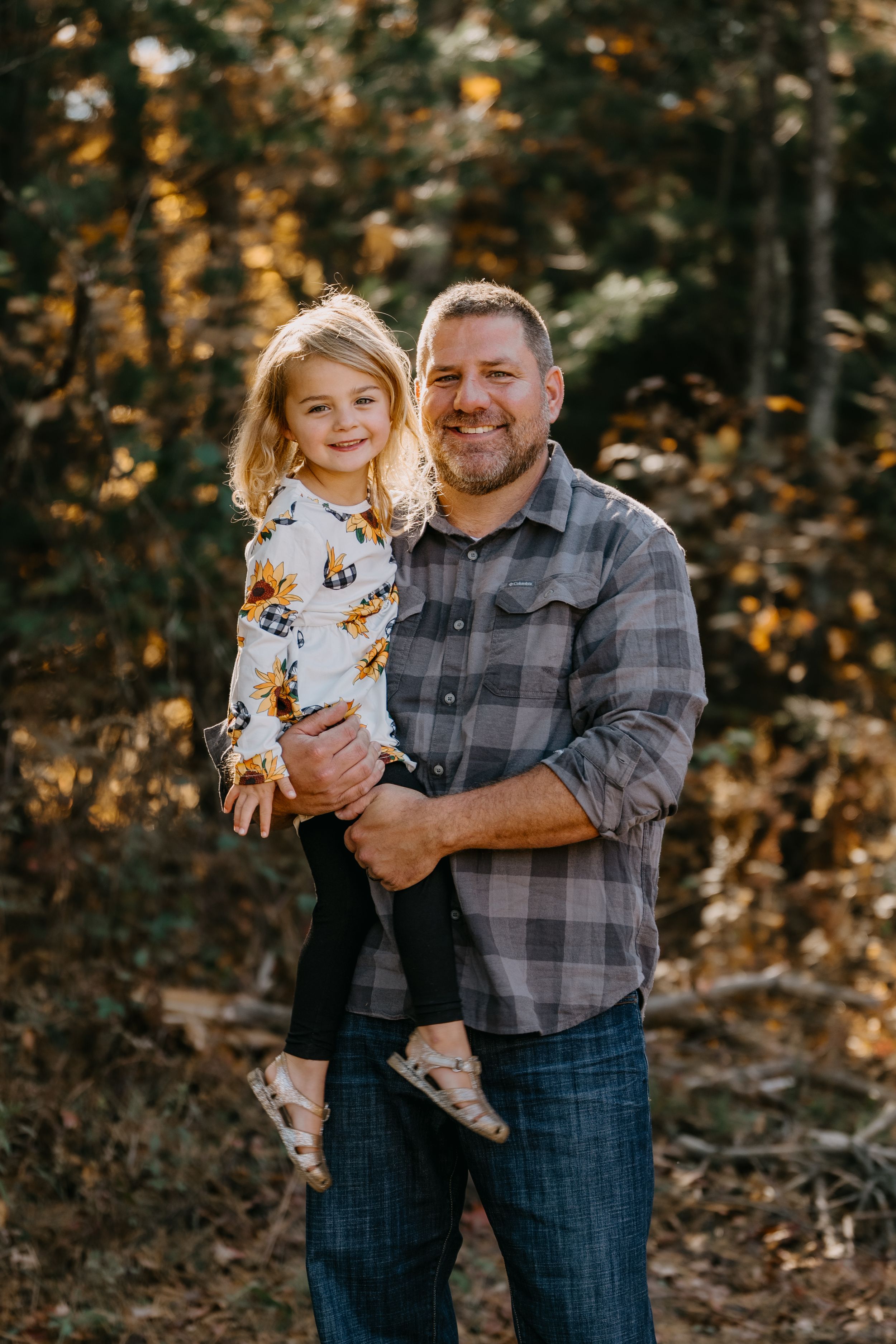 Outdoor portrait in autumn woods with golden light filtering through trees behind figures in plaid and floral.