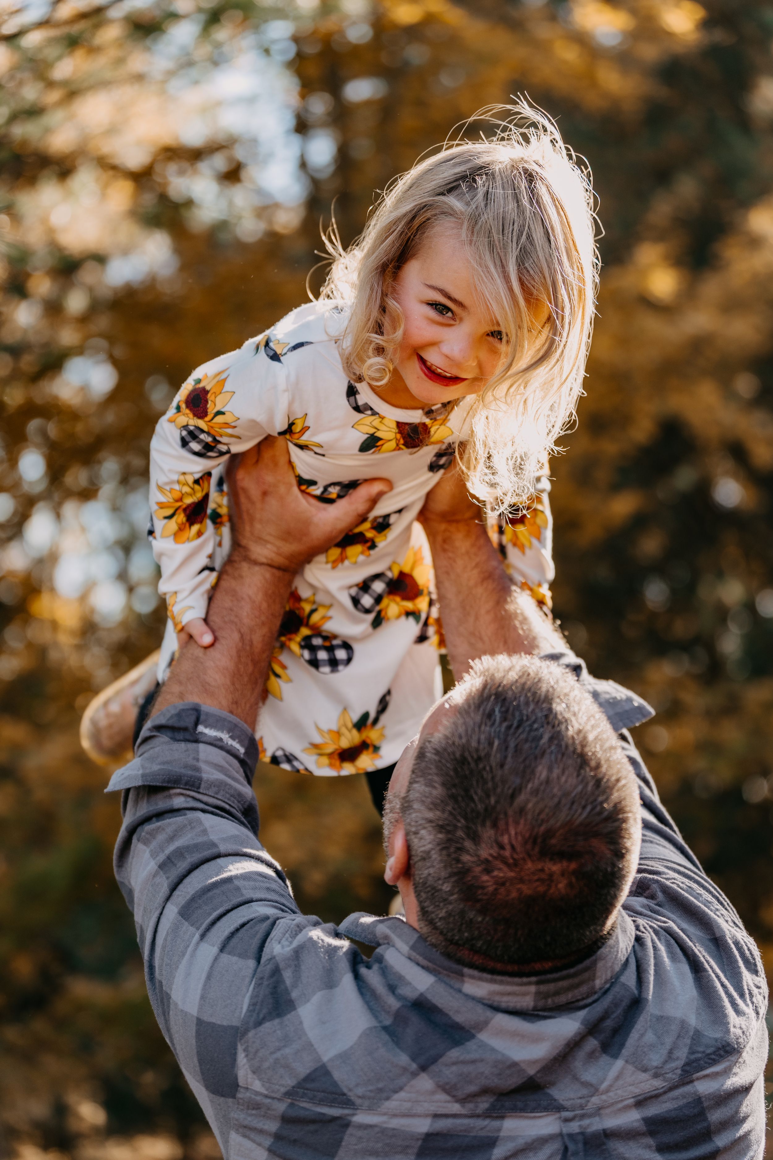 A child wearing a floral print shirt is lifted up by someone in a plaid shirt on a sunny autumn day in the woods.