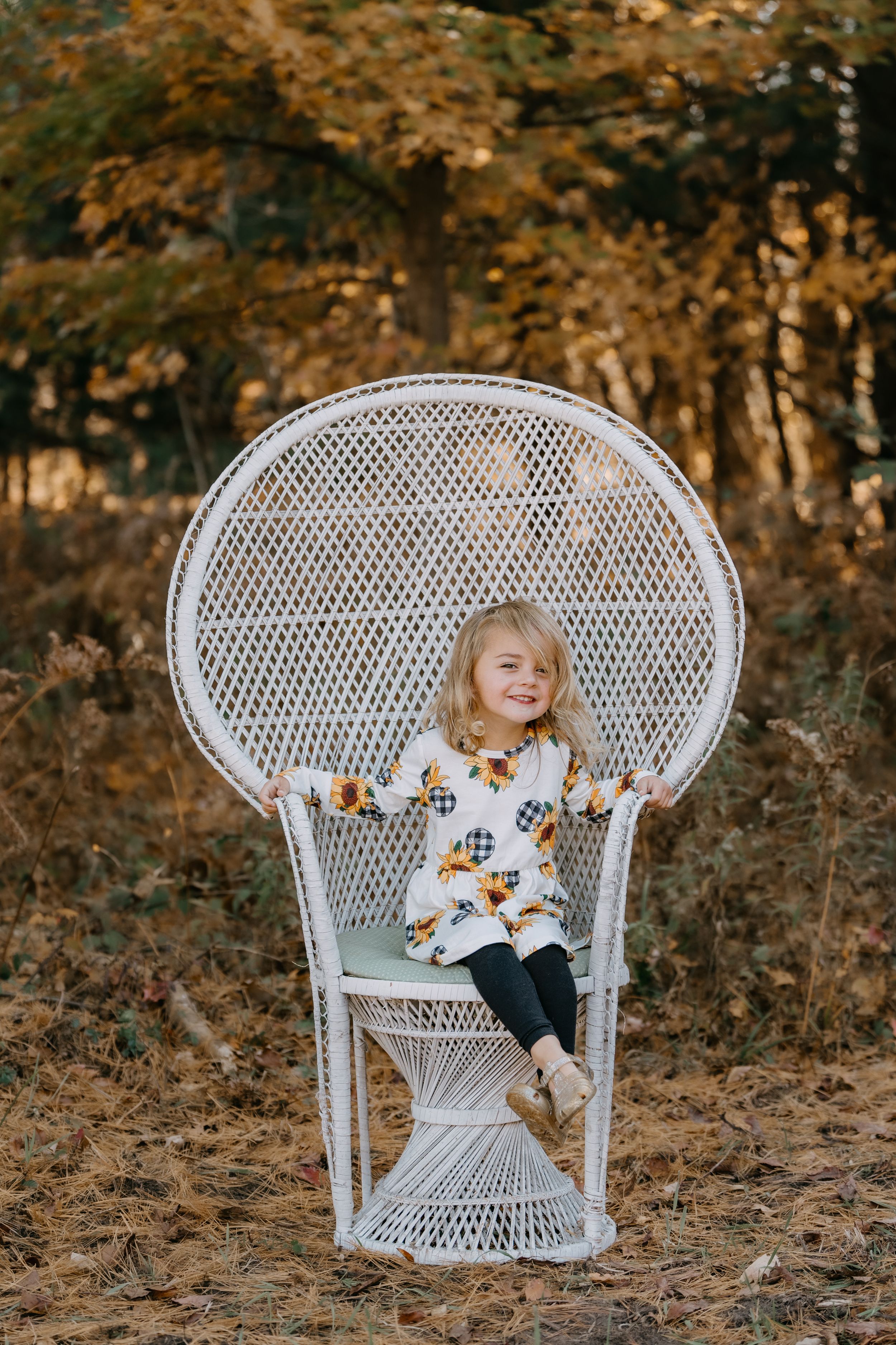 A toddler sits happily in a large white wicker peacock chair outdoors with autumn foliage in the background.