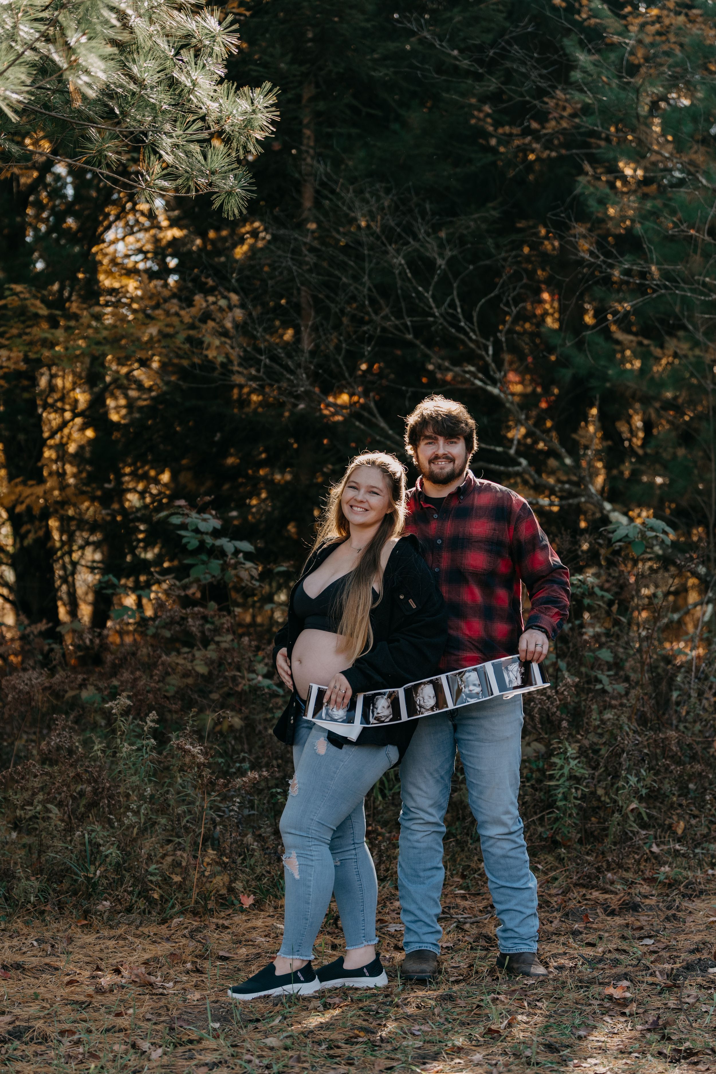Couple in autumn forest holding ultrasound photos while expecting their first child.
