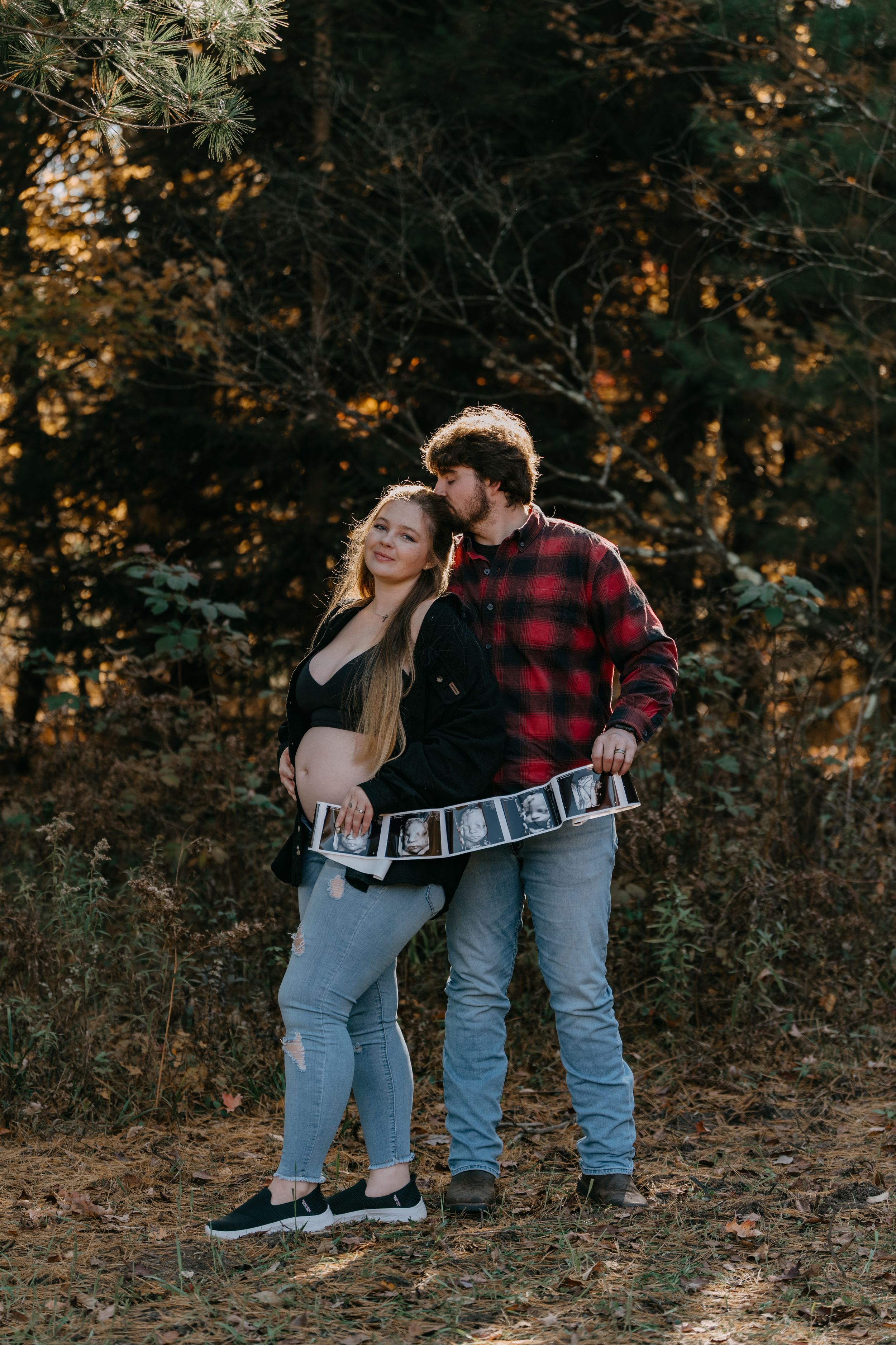 Couple holds ultrasound strip in autumn forest during maternity photoshoot with warm golden lighting.
