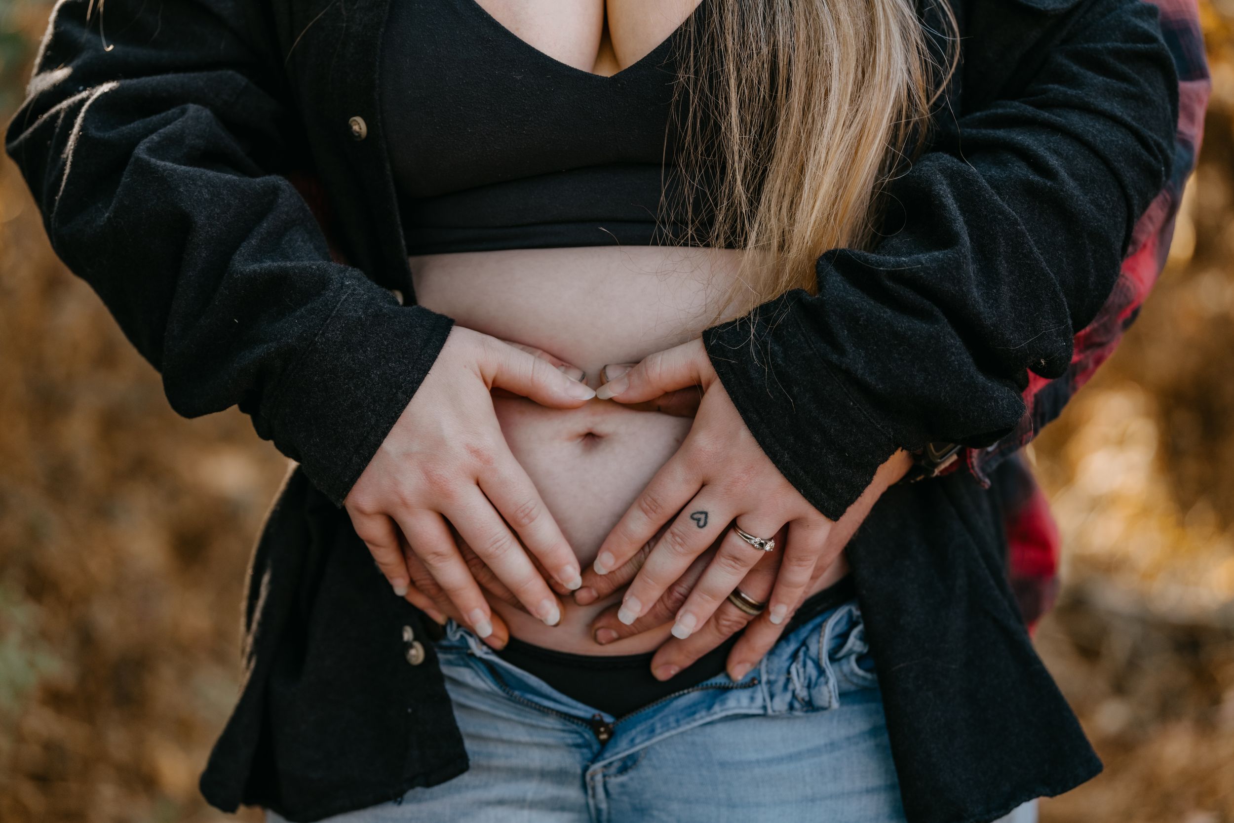 Hands form a heart shape over a woman's belly while wearing a black jacket and jeans.