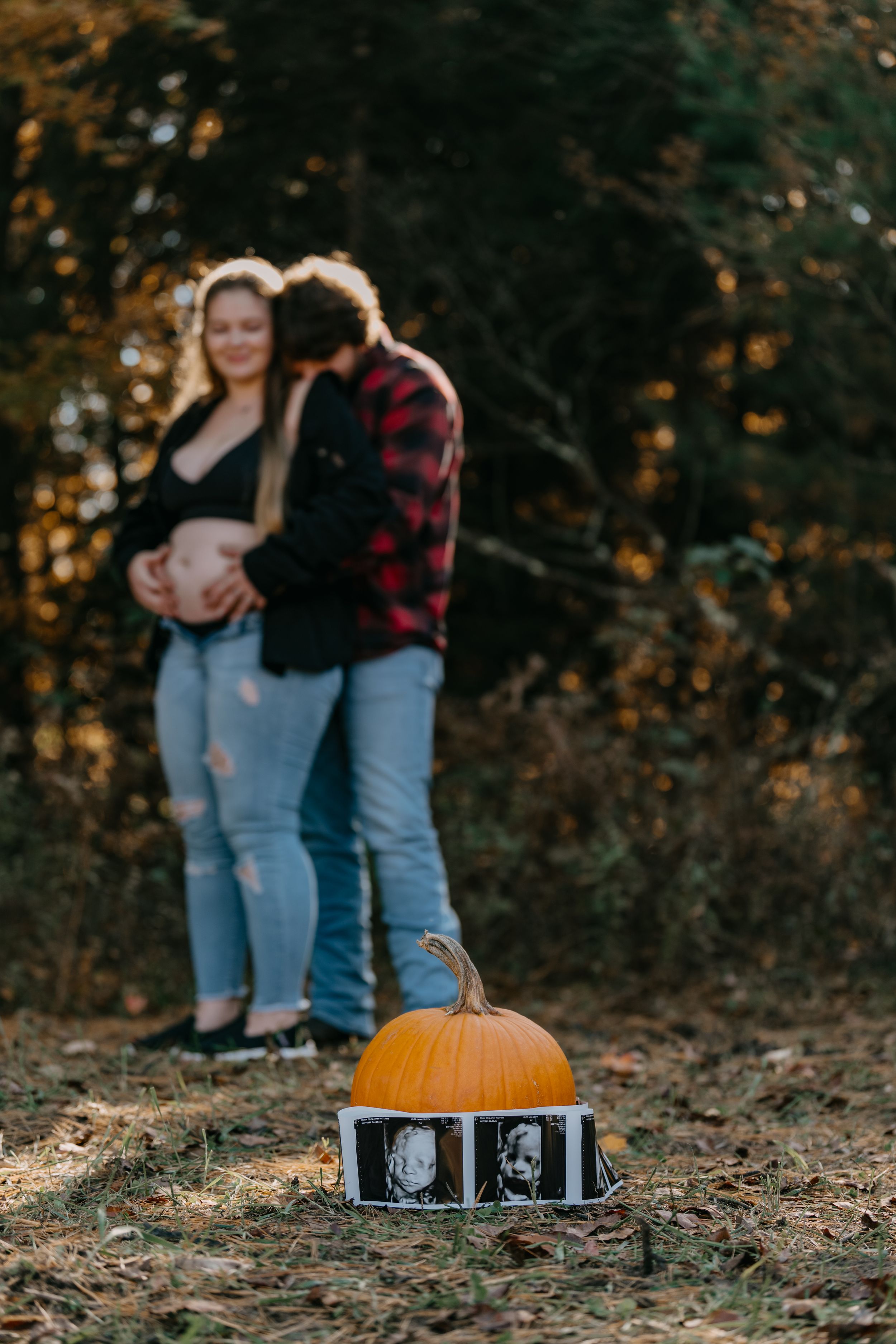 Autumn maternity photo with pumpkin and ultrasound strip displayed in foreground of wooded setting.