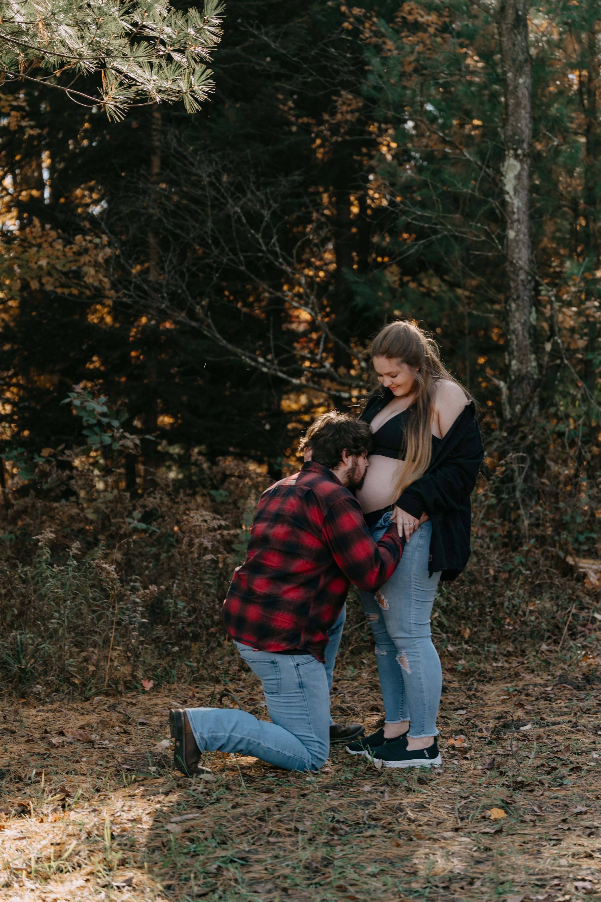 Expecting couple shares intimate forest maternity moment with person kneeling.