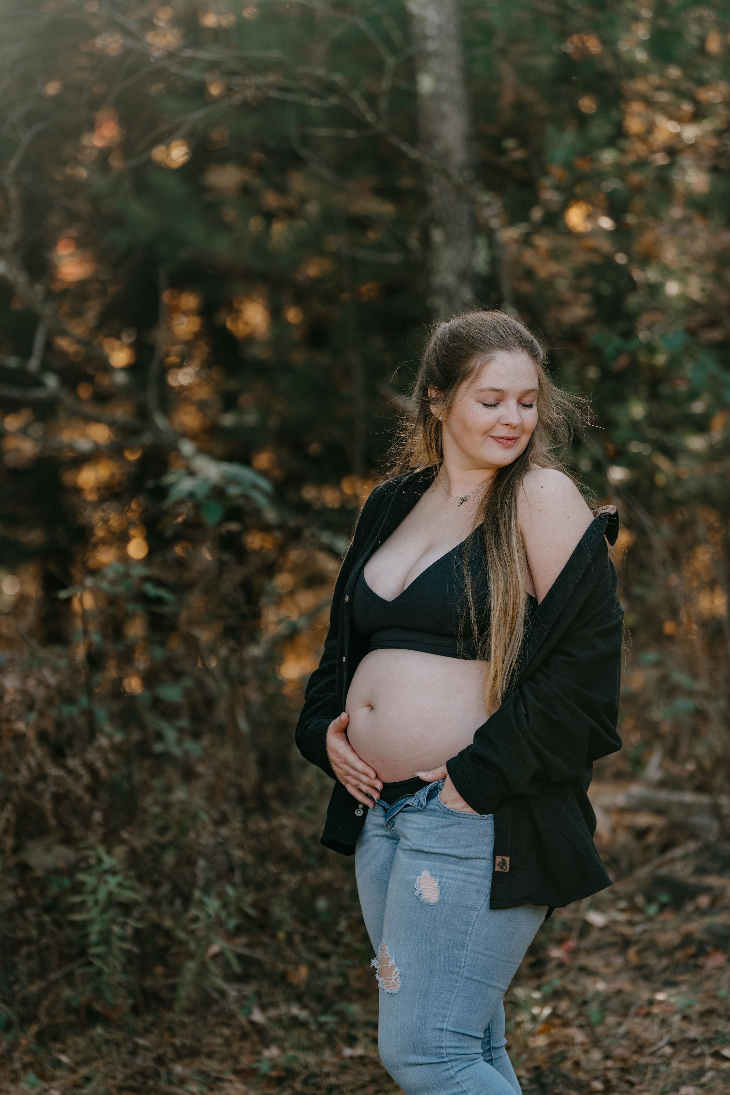 Pregnant woman in denim and black jacket smiles while cradling baby bump in autumn woods.