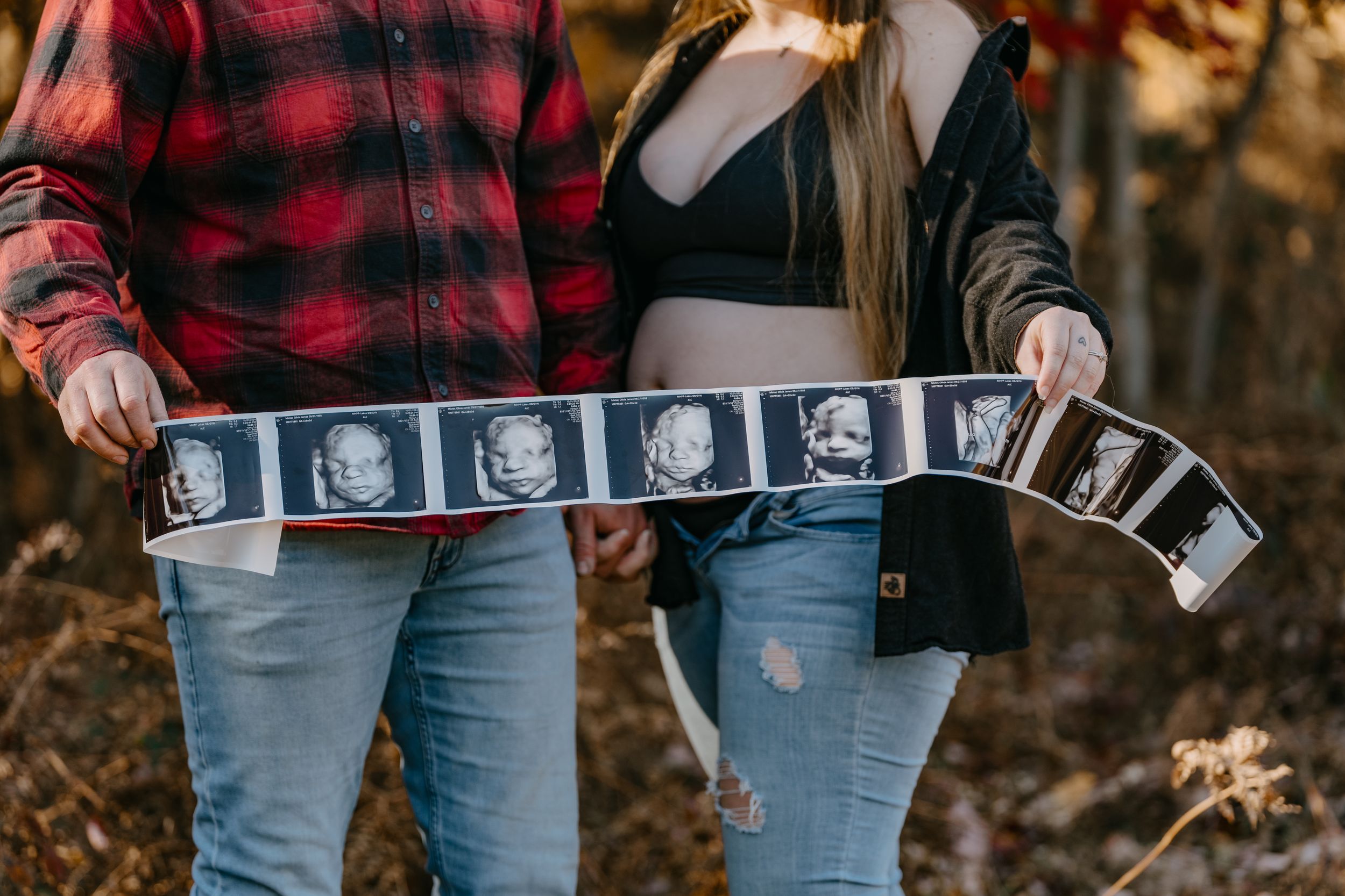 A couple holding a strip of ultrasound photos while wearing plaid and denim in an outdoor setting.