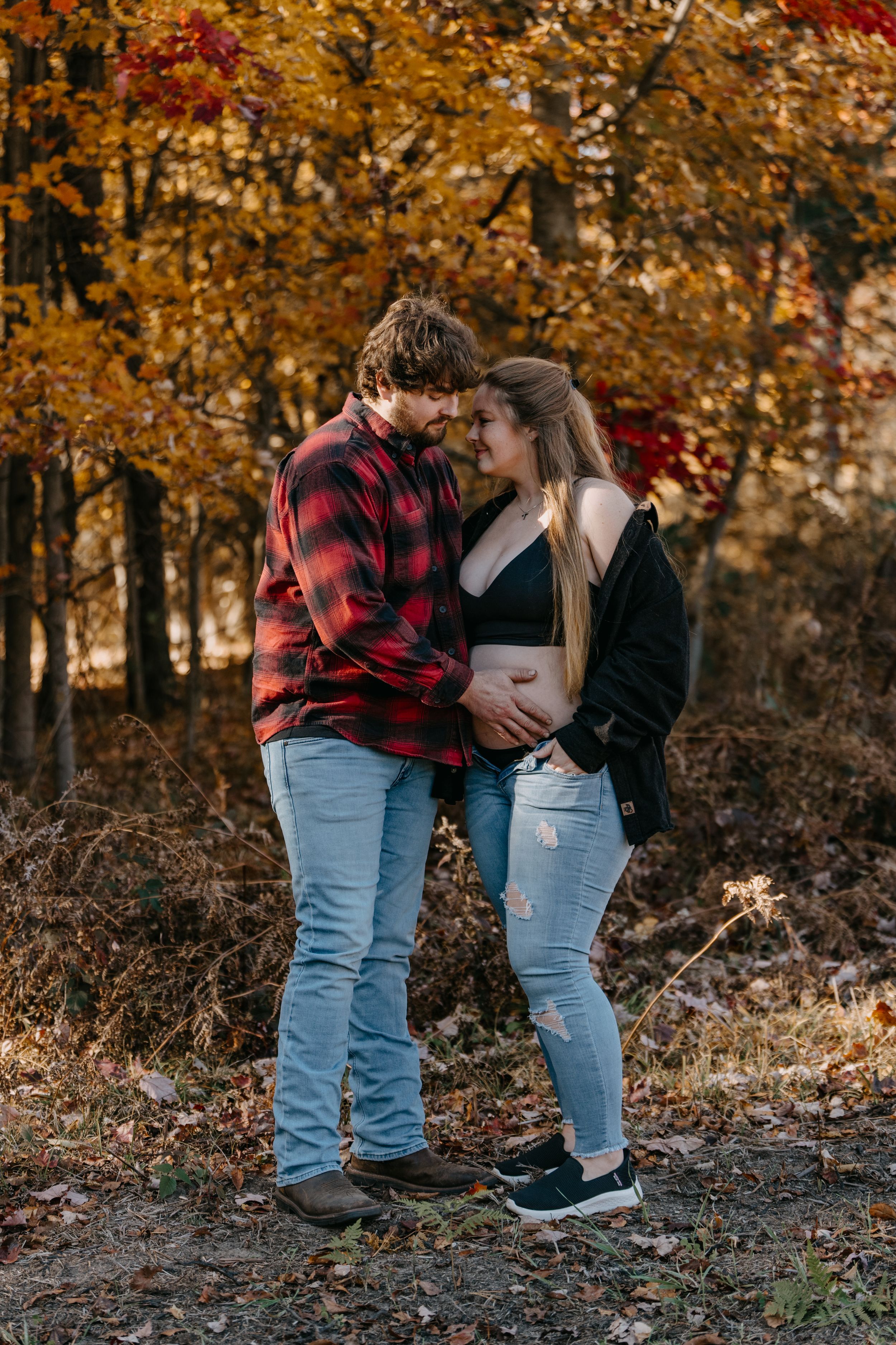 An expectant couple embraces in front of golden autumn trees wearing matching casual attire.