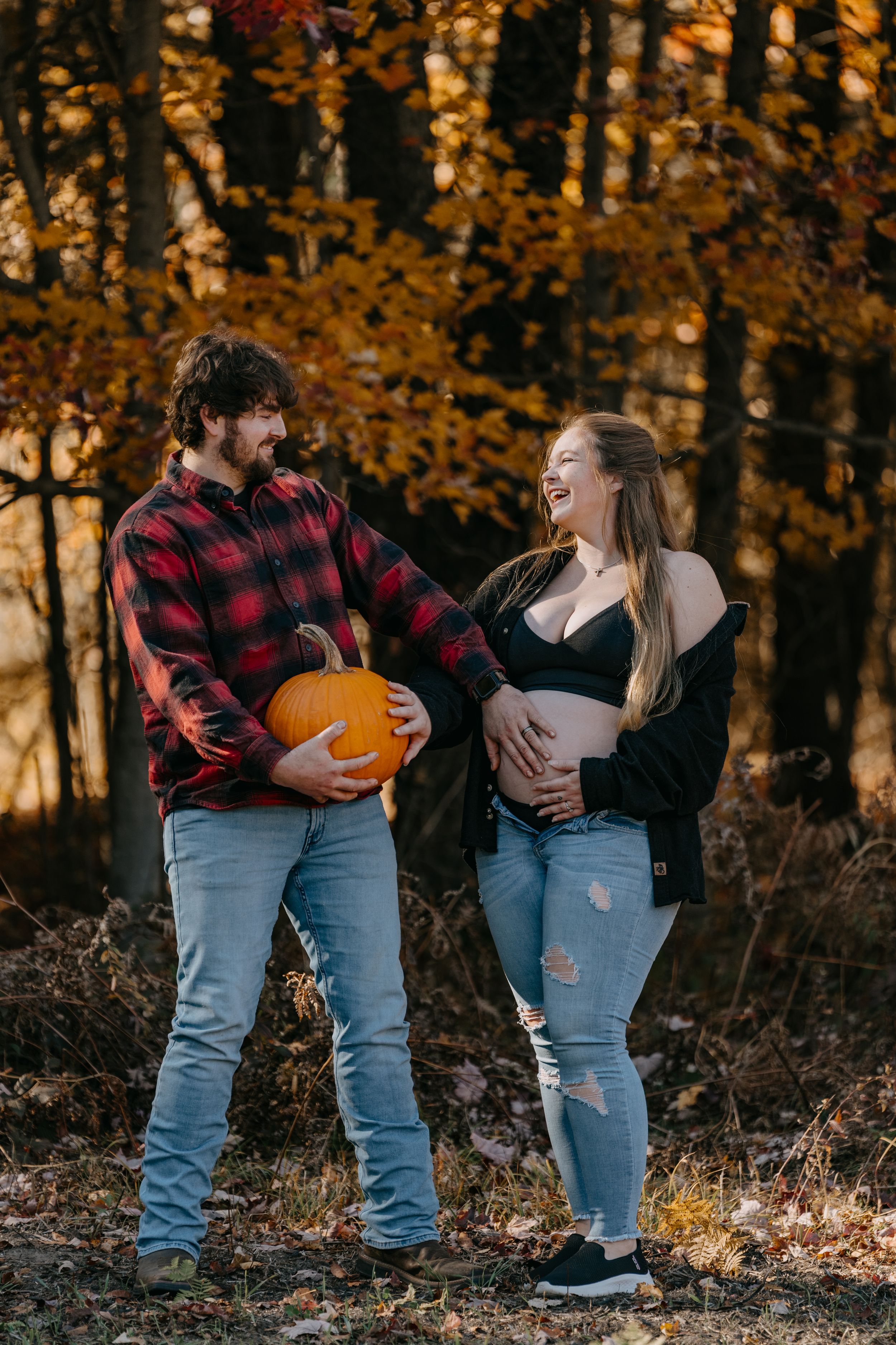 Expecting couple shares joyful moment with pumpkin in autumn forest during maternity photoshoot.
