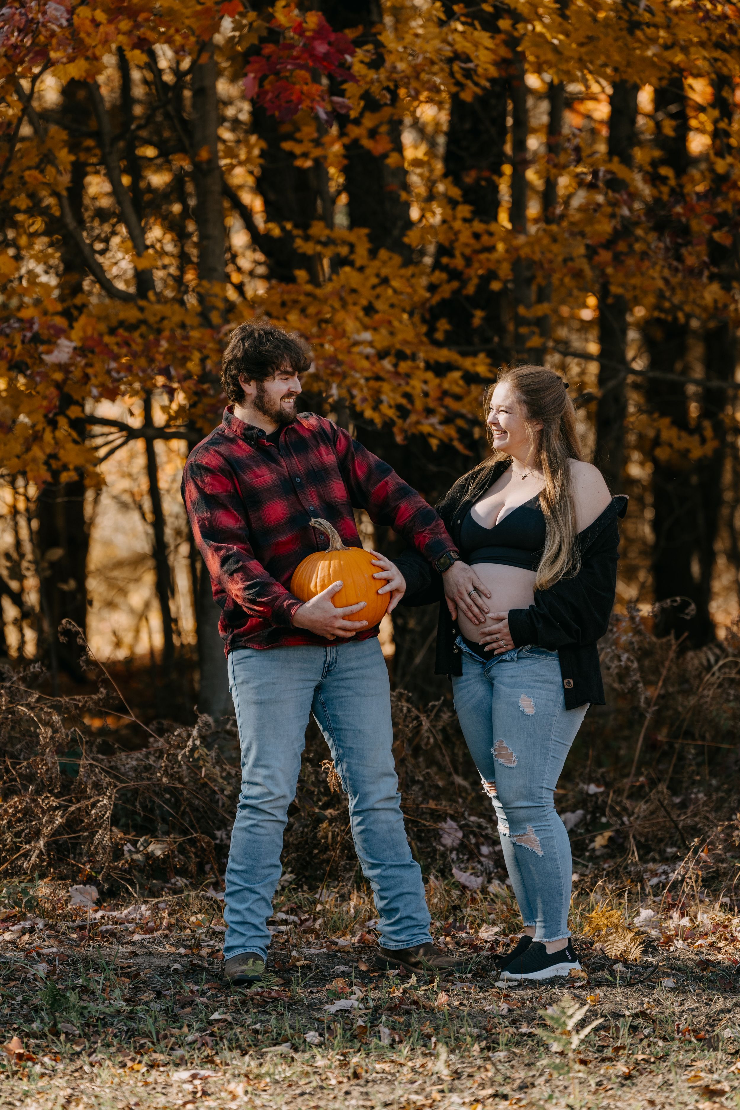 A couple in casual wear poses with a pumpkin and belly bump against an autumn forest backdrop.