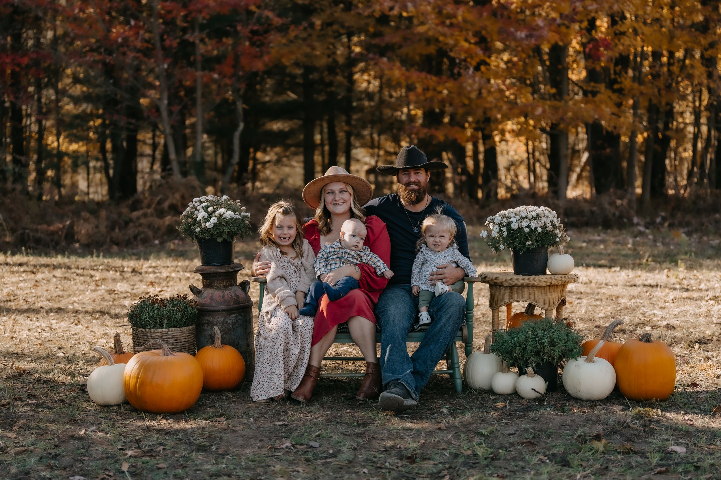 Fall family photo setup in a wooded area with pumpkins, white chairs, and autumn decor.