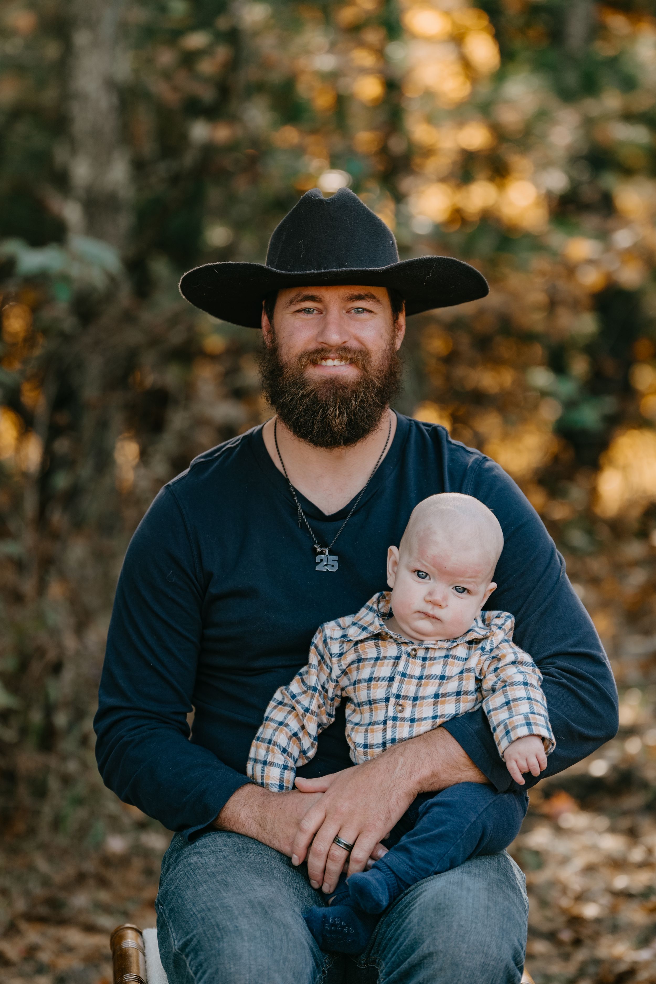 A father in a black cowboy hat holding a baby in plaid clothing during an outdoor autumn photoshoot.