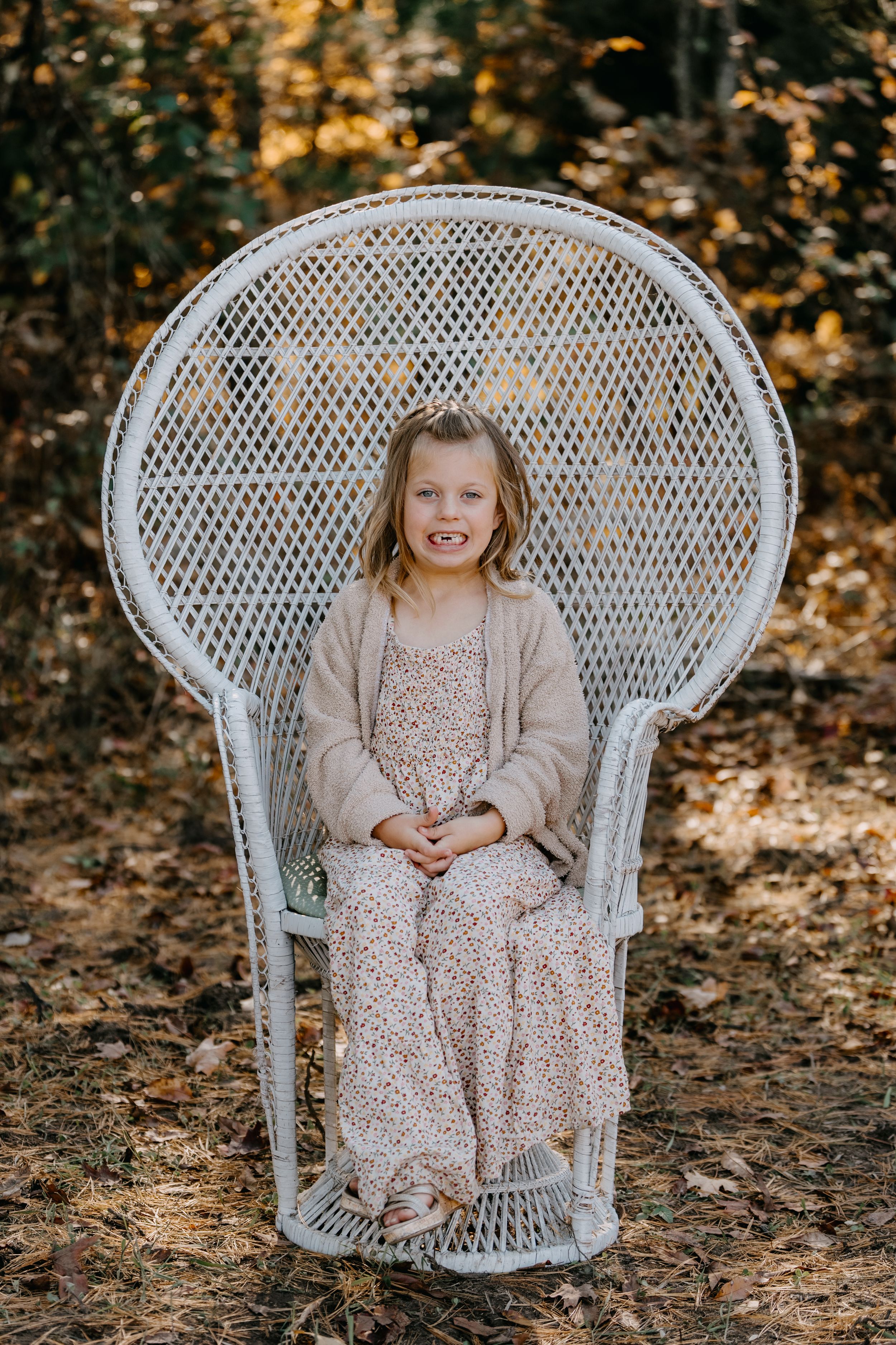 A white wicker peacock chair set up for family photos in a forest with fall foliage.