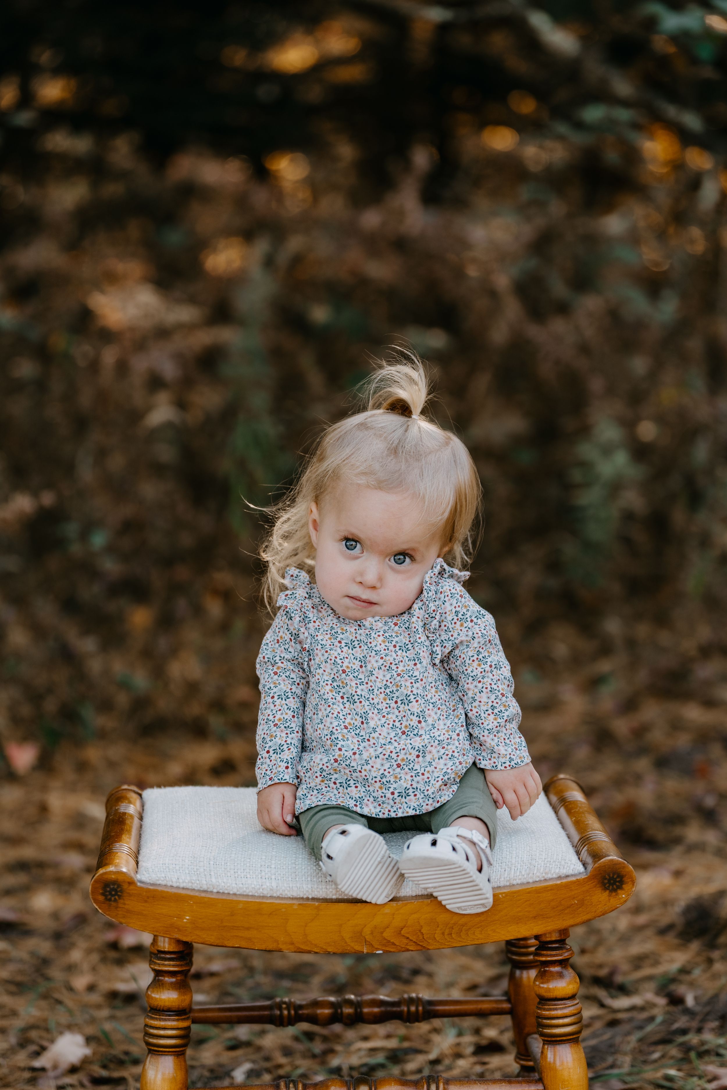 A small child sits on a wooden chair during an autumn outdoor photoshoot.