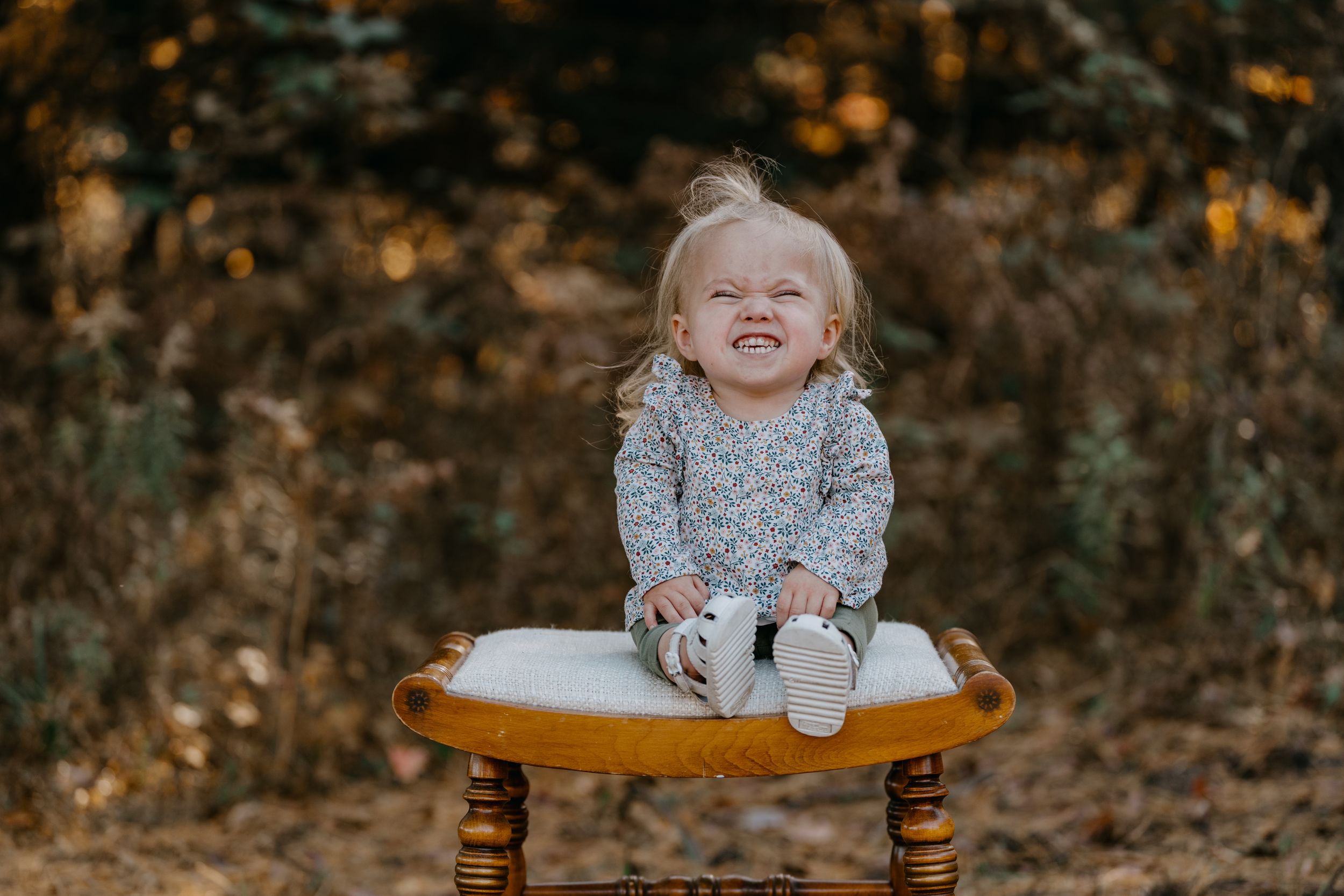 Small child wearing grey sweater sits joyfully on wooden chair in autumn outdoor setting.
