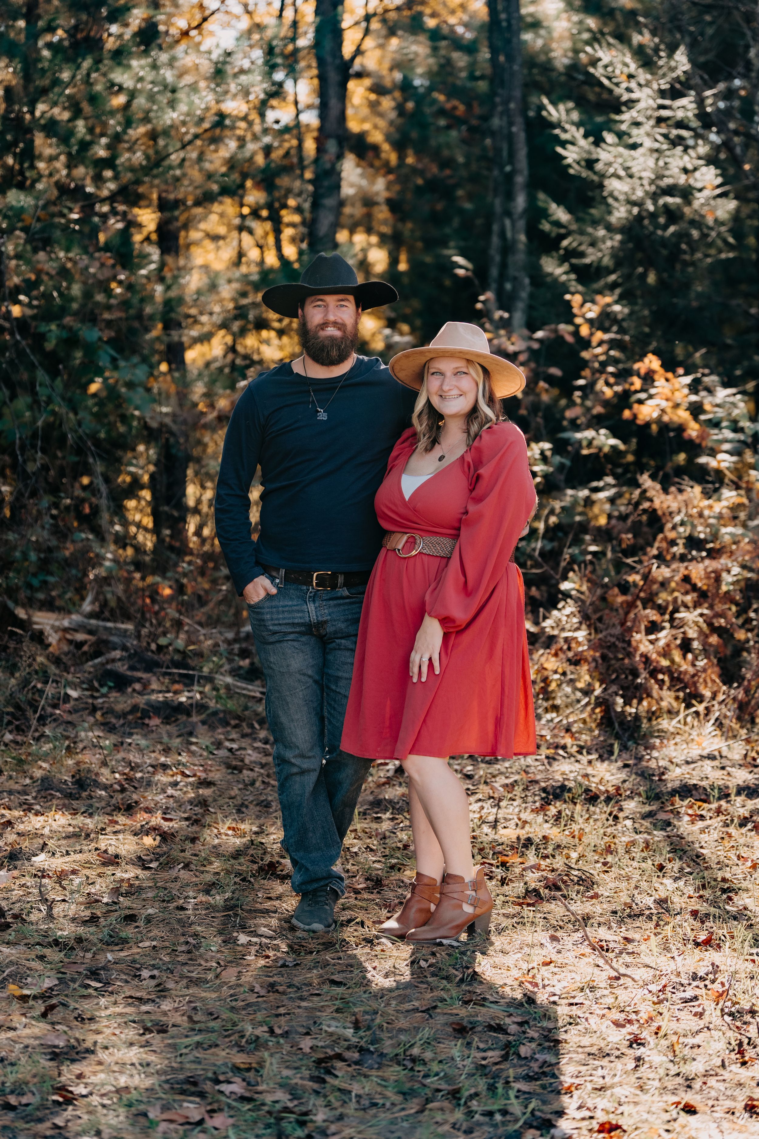 A couple in casual attire poses together against a sunlit autumn forest backdrop.