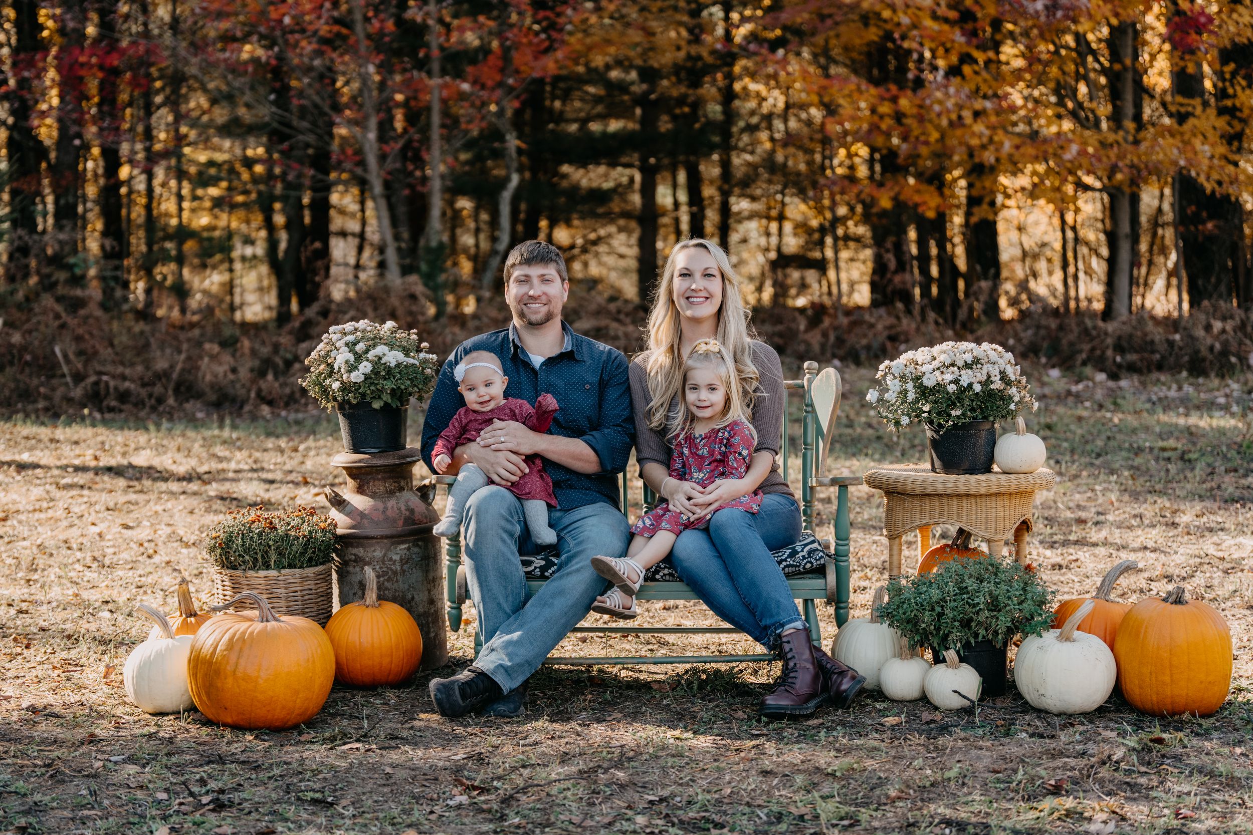 A family sits together outdoors on a fall day with pumpkins and autumn foliage in the background.