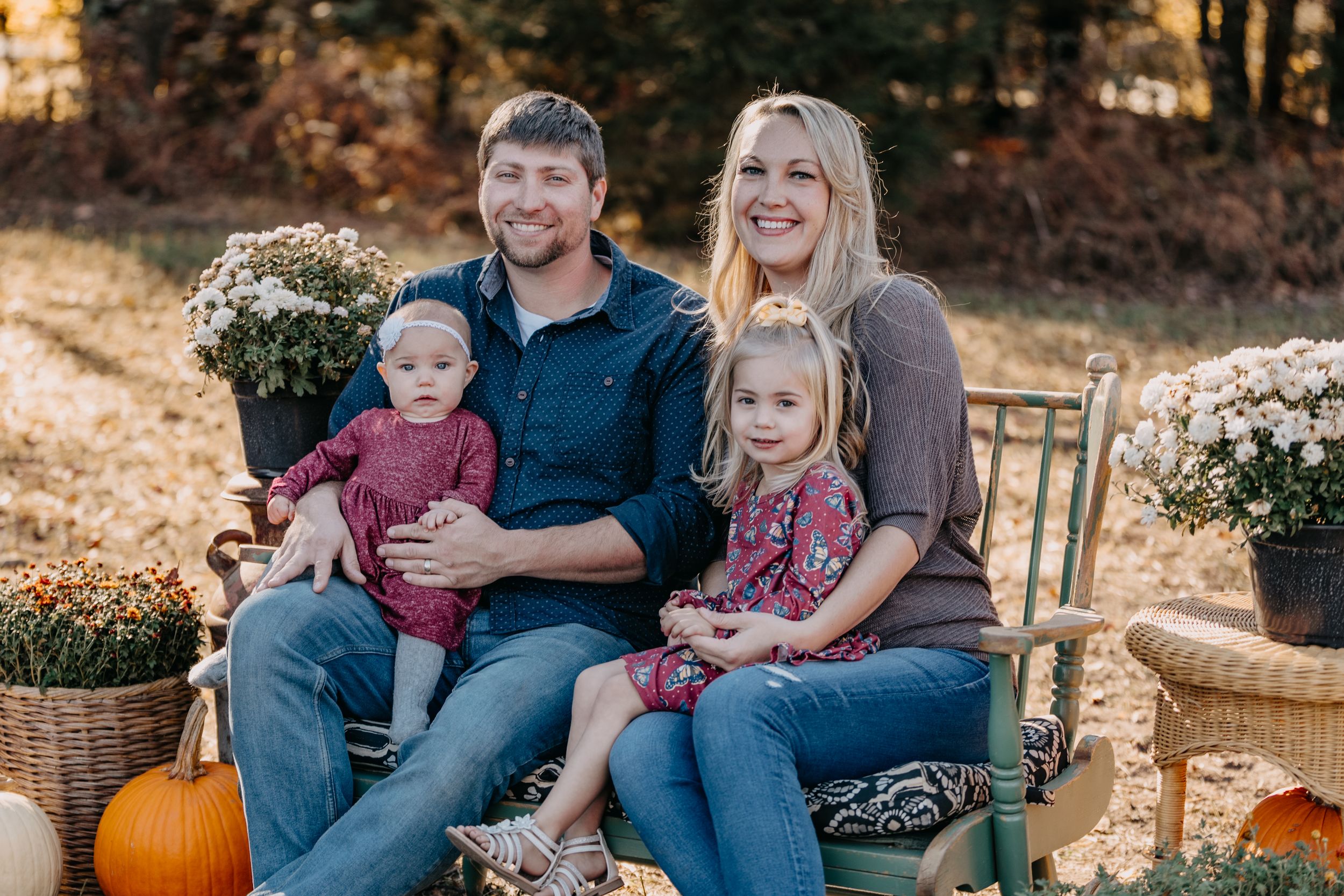 A group sits on an outdoor chair surrounded by fall decorations including pumpkins and mums in golden autumn light.