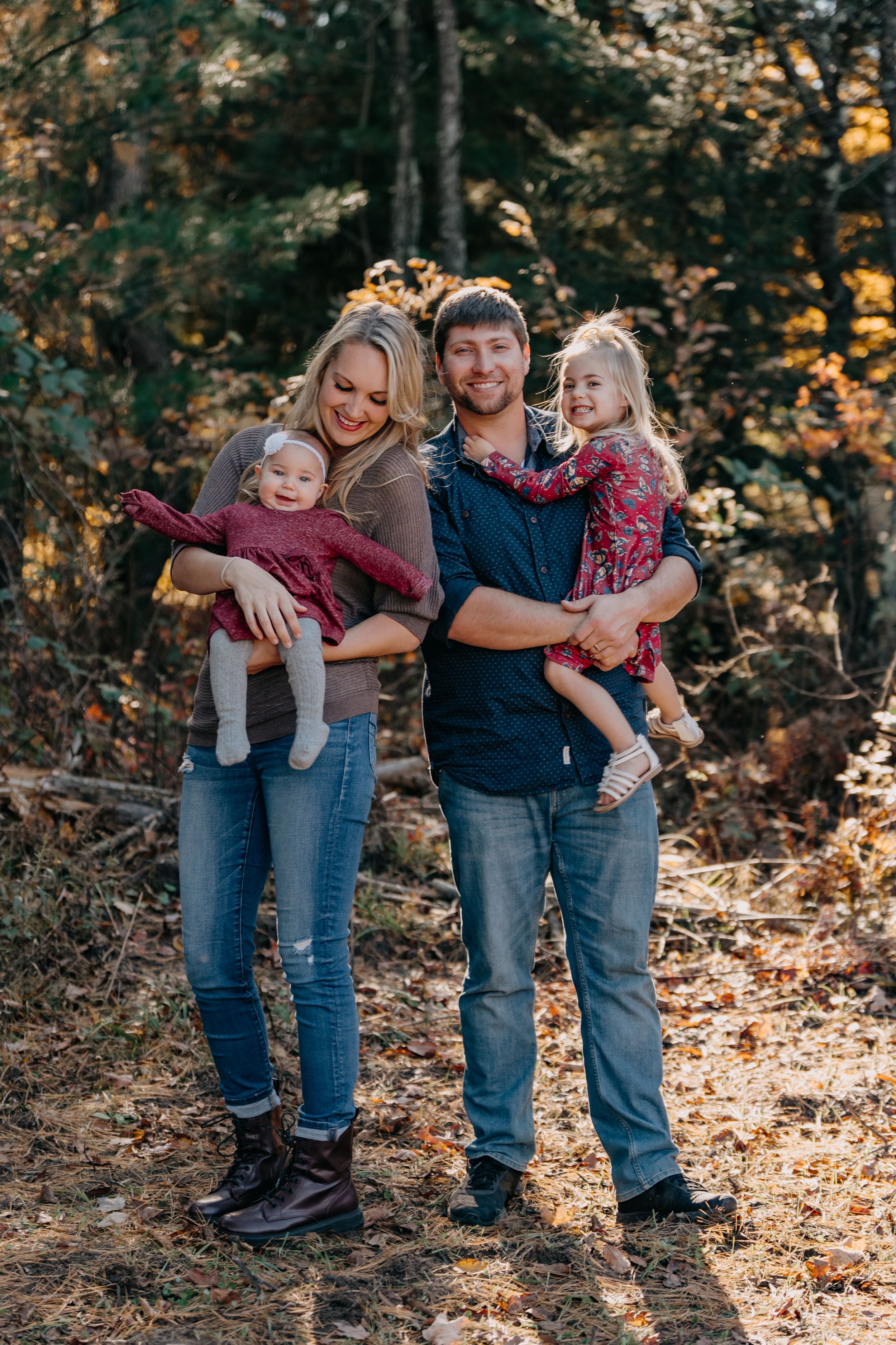 Family in casual attire posing together during autumn photoshoot in a wooded forest setting.