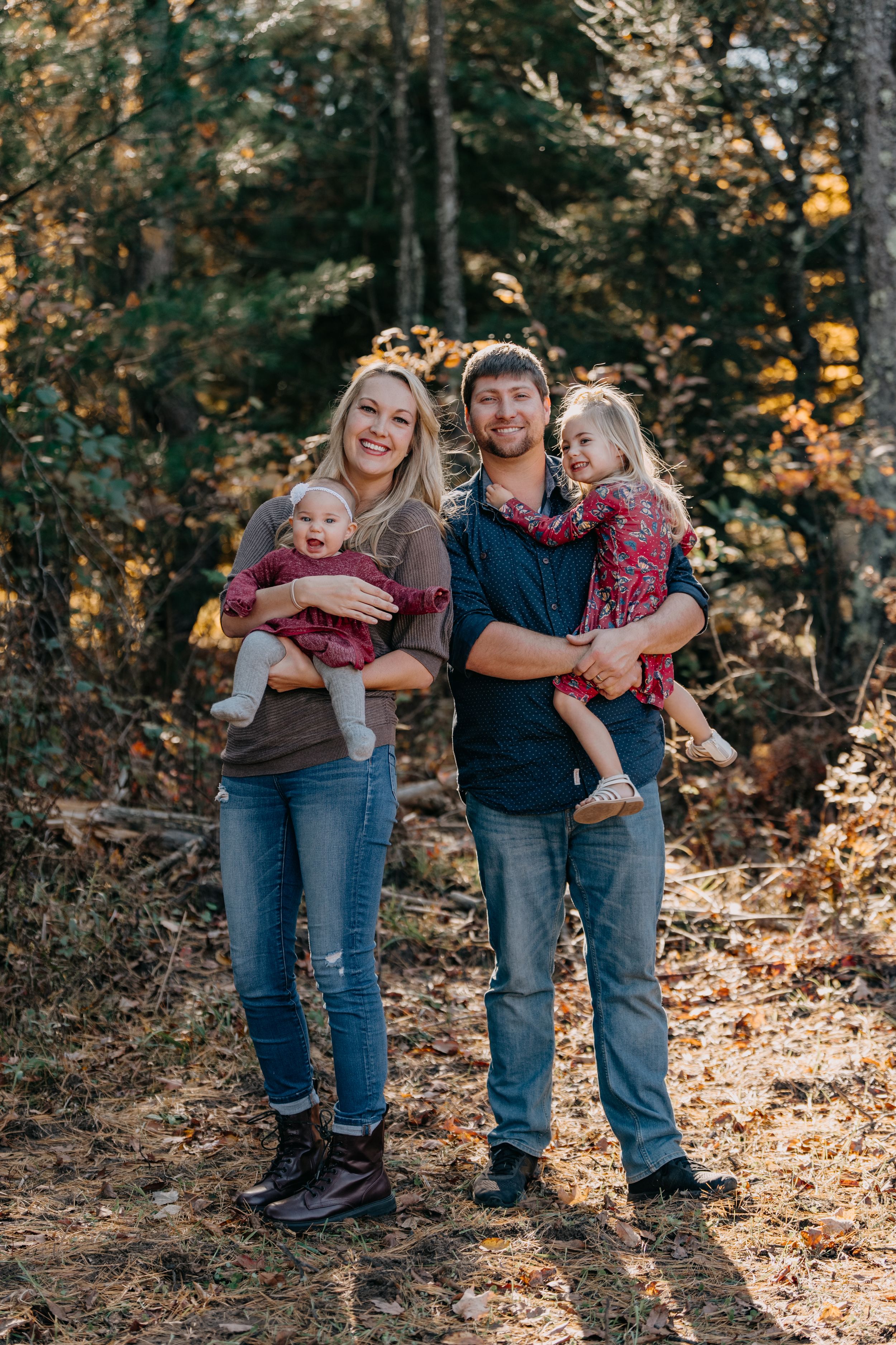 A group stands together on a wooded autumn trail wearing coordinating burgundy and denim outfits in golden sunlight.