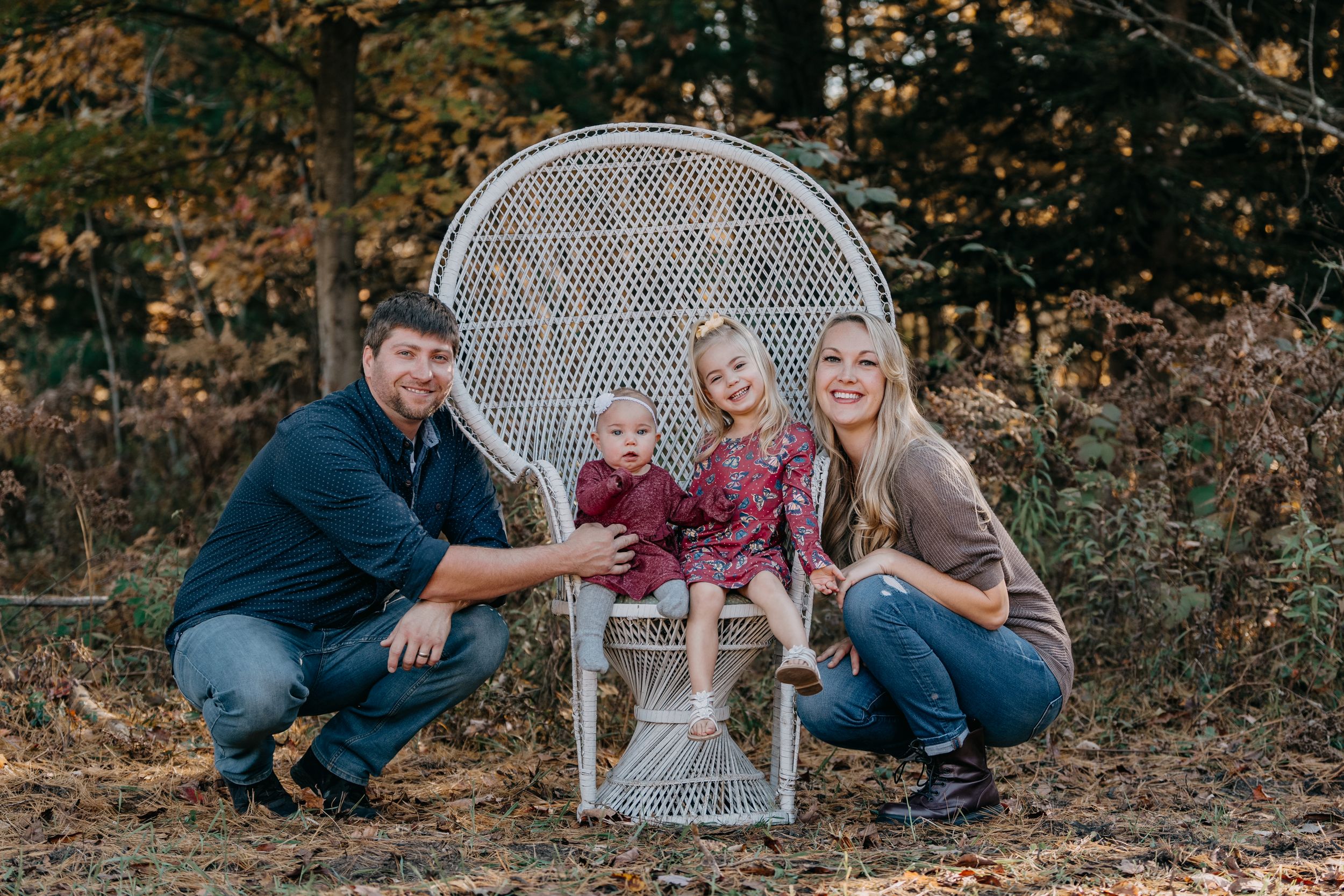 Autumn outdoor family portrait with wicker peacock chair, burgundy clothing, and a wooded background.