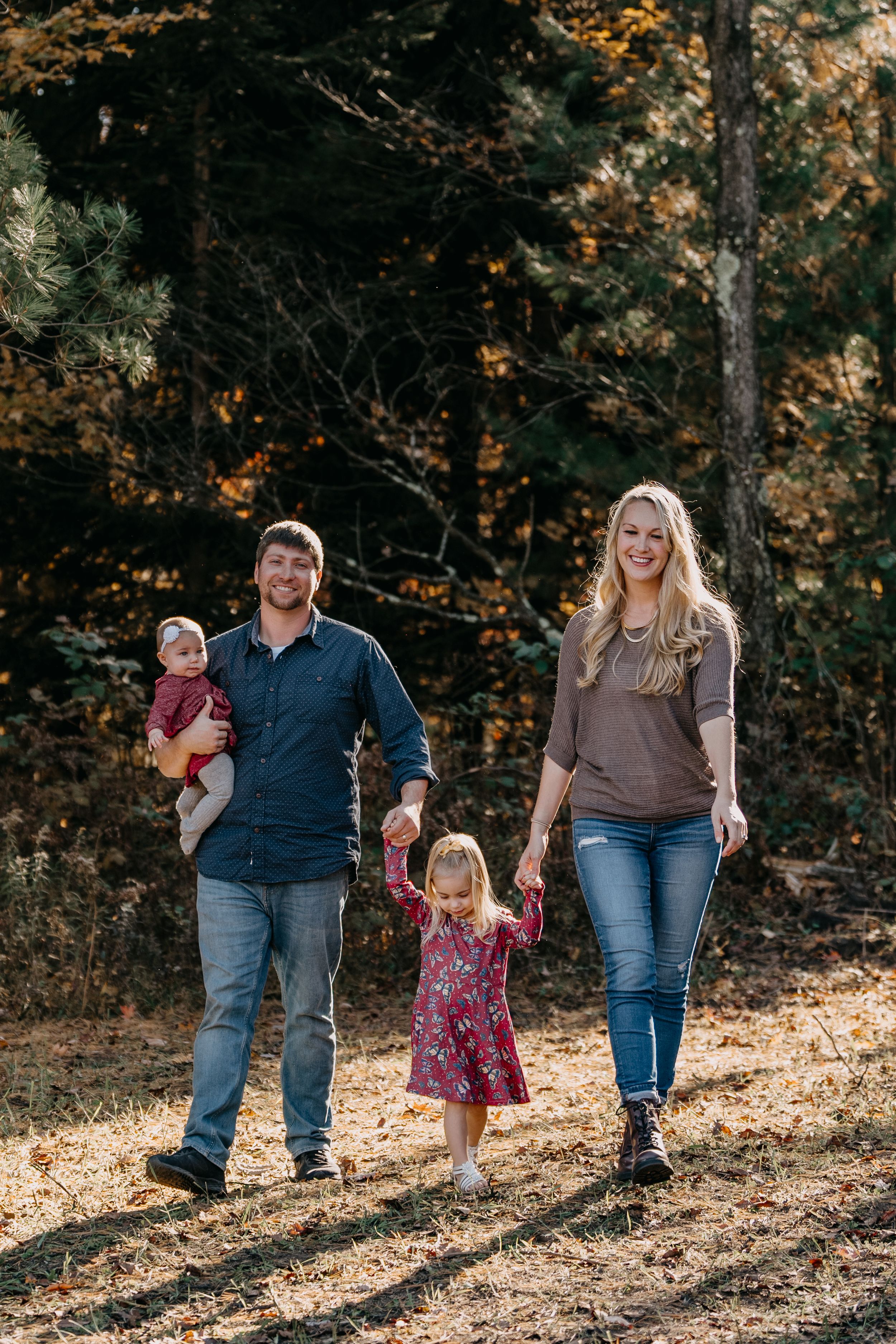 A family walks together on a wooded trail during autumn with fallen leaves and golden sunlight filtering through trees.