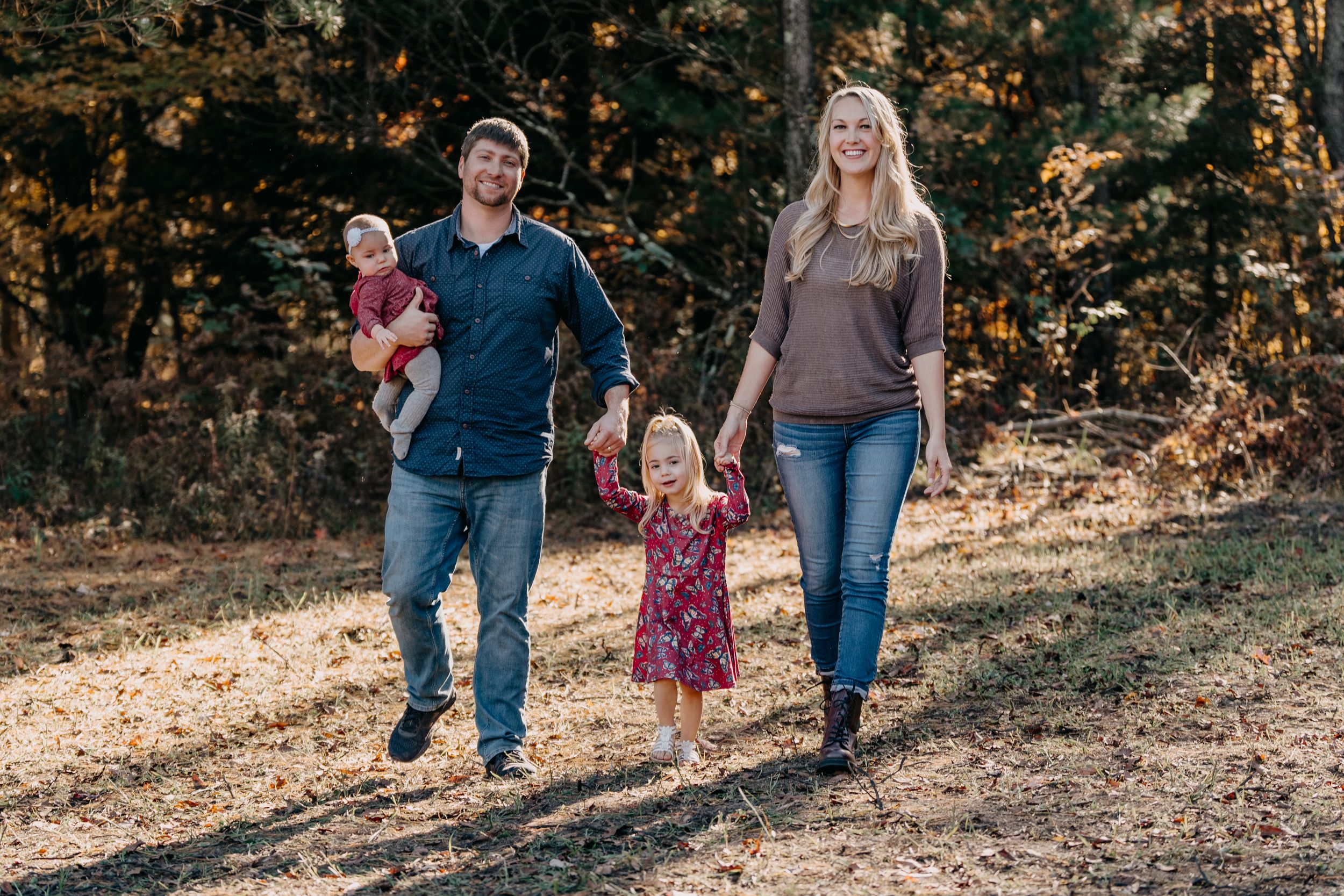 A family walks together on a wooded path during a fall photo session with golden sunlight filtering through trees.