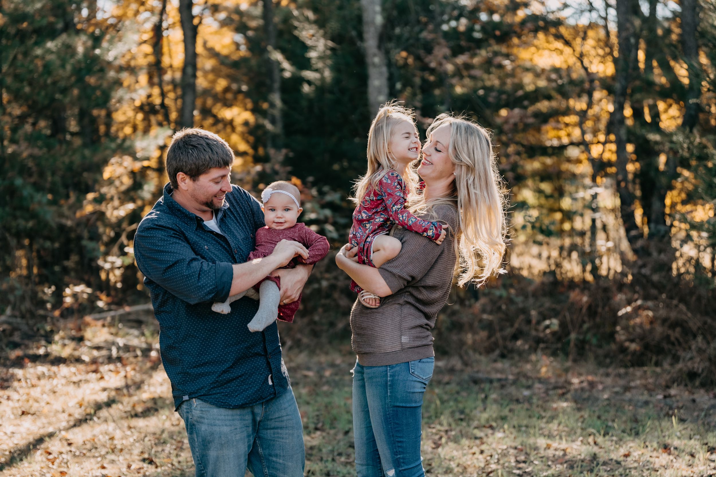 Family enjoys a playful autumn photoshoot together in a sun-dappled wooded area during golden hour.