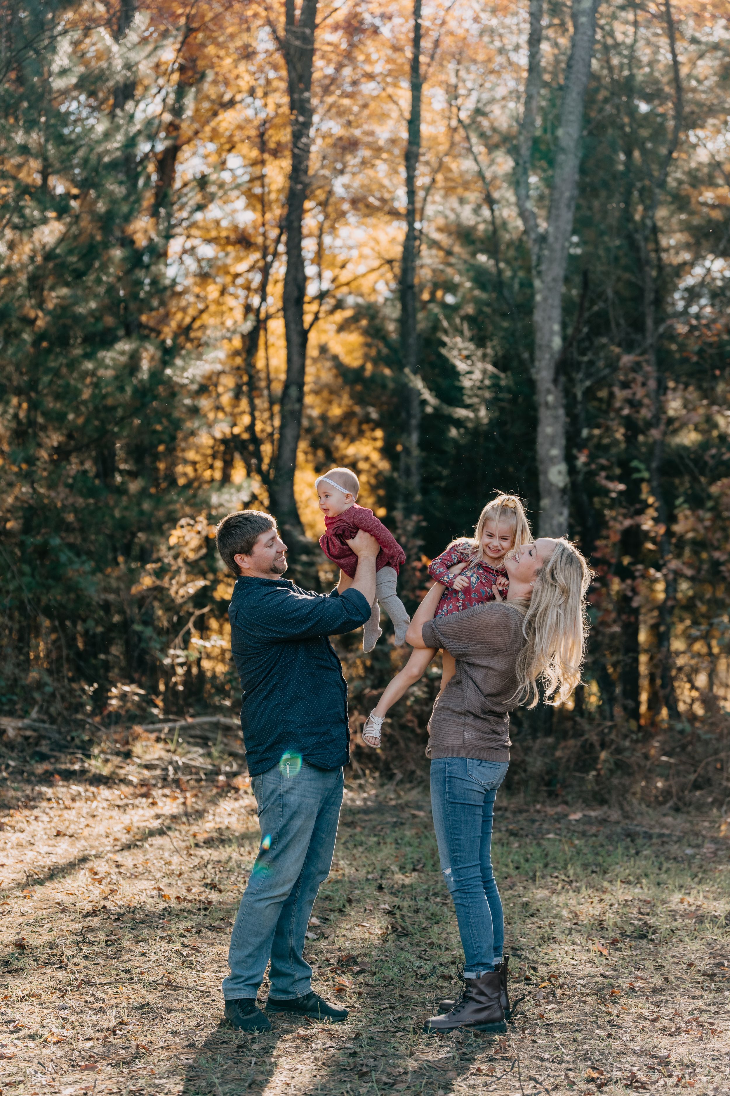 A couple plays with their children in a sunlit autumn forest during a family photoshoot.