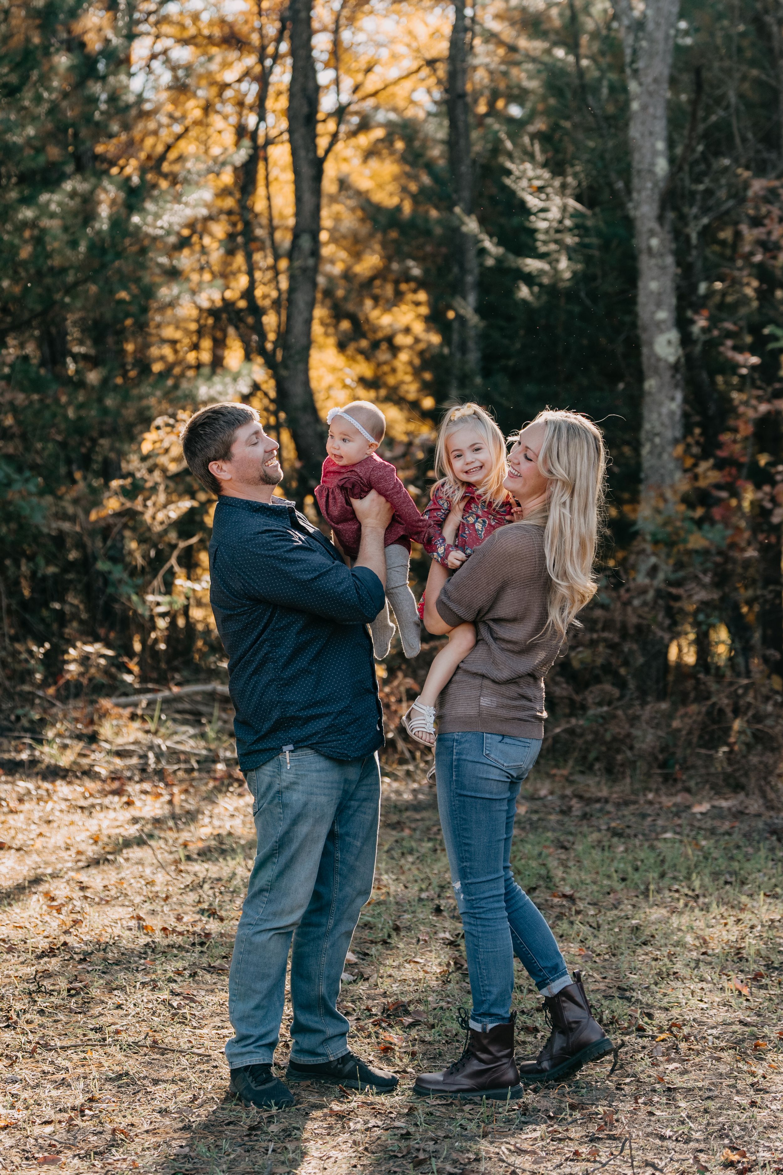 A family shares joyful moments together in an autumn forest setting with golden sunlight filtering through the trees.