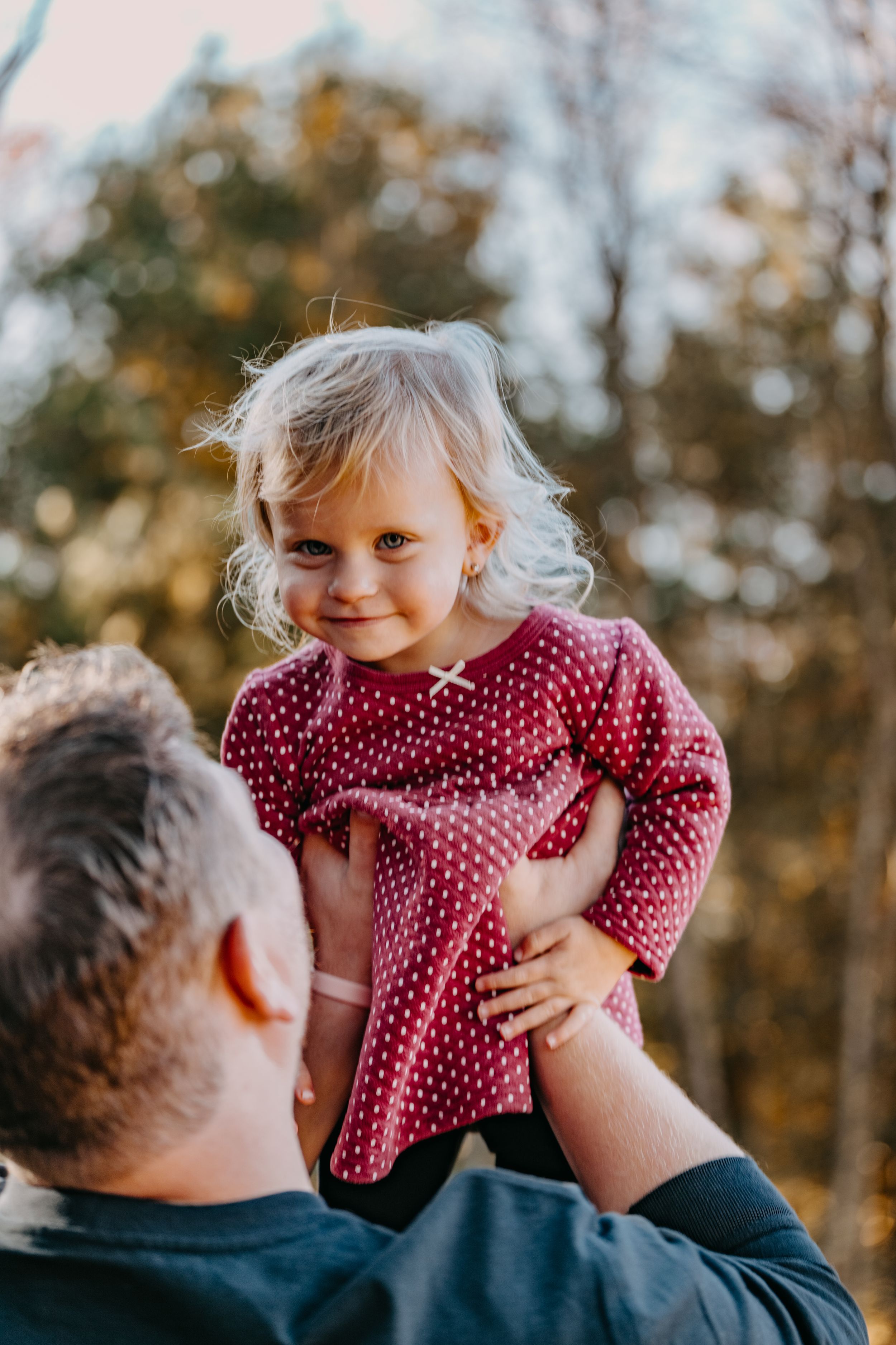 Small child in red polka dot dress being lifted up by adult wearing navy shirt on autumn afternoon.