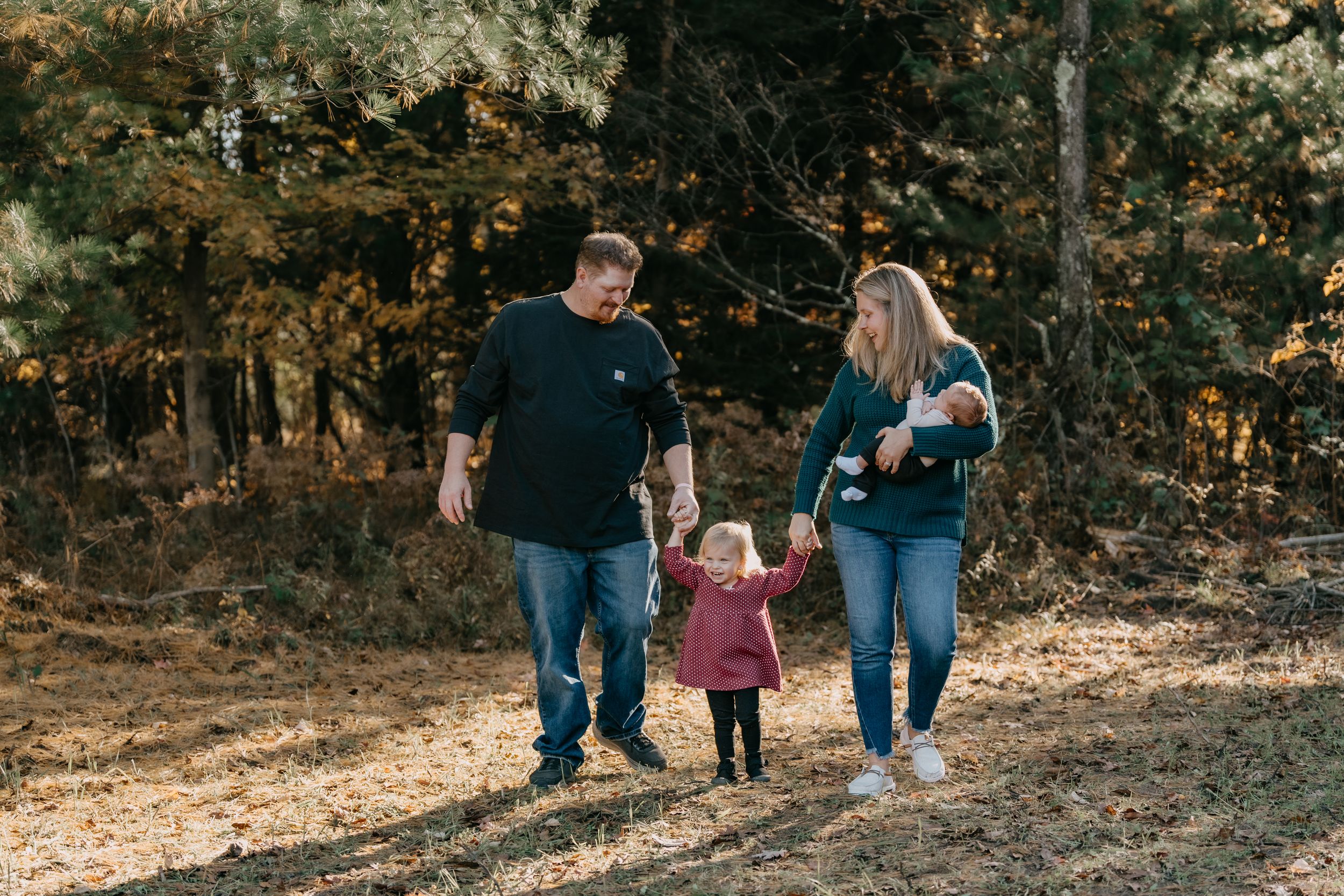 A family walking through a sunlit autumn forest holding hands along a leaf-covered path.