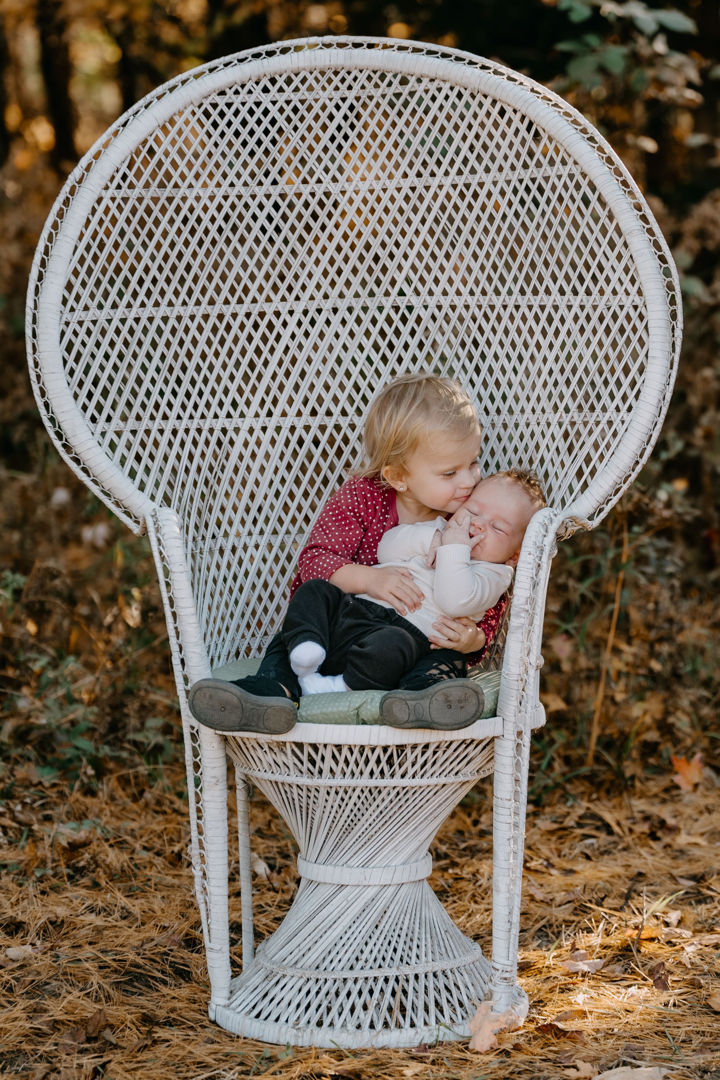 White wicker peacock chair holds two small children together in a serene outdoor autumn setting.