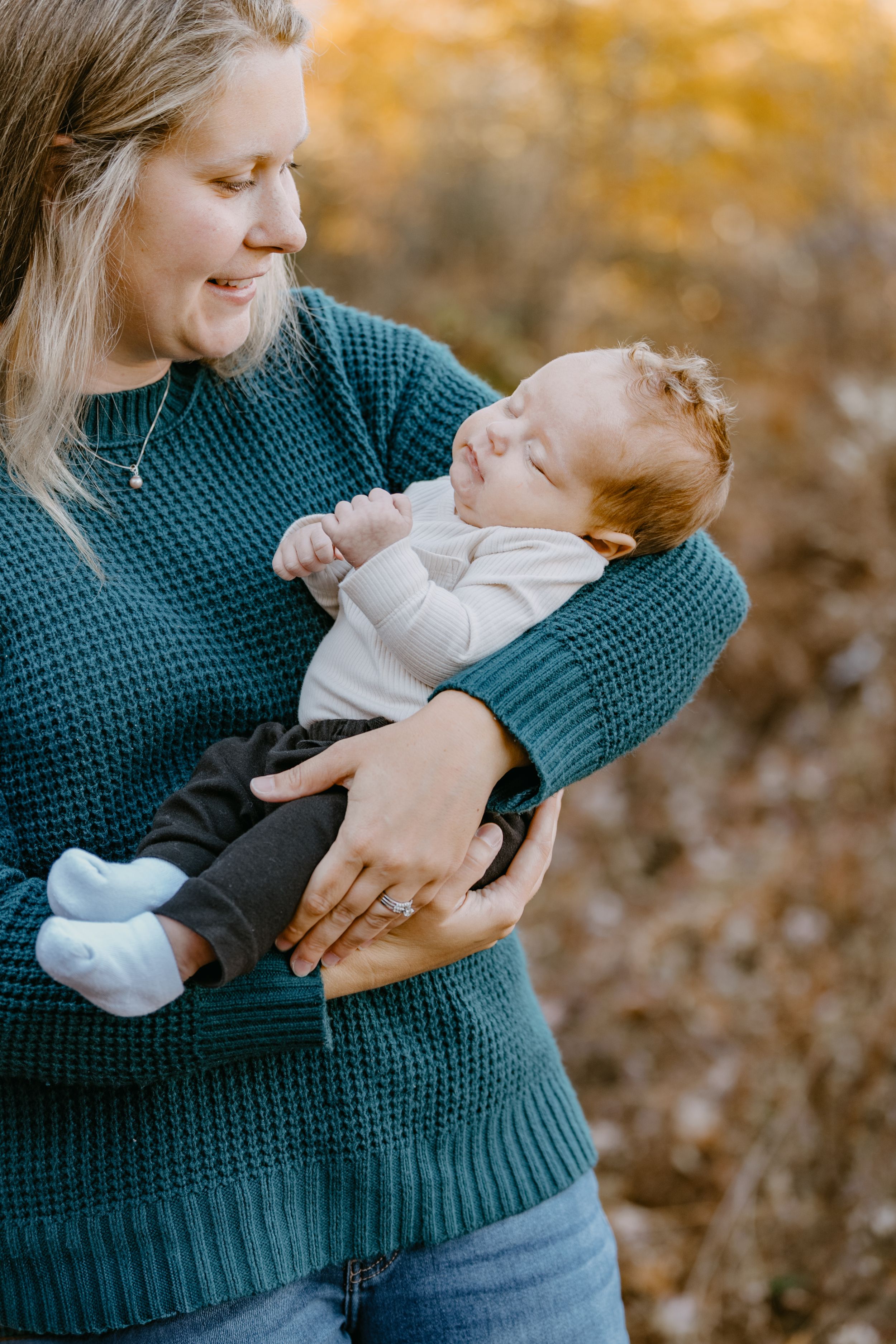 A caregiver in a teal knitted sweater holds a sleeping infant wearing white and gray in an autumn outdoor setting.