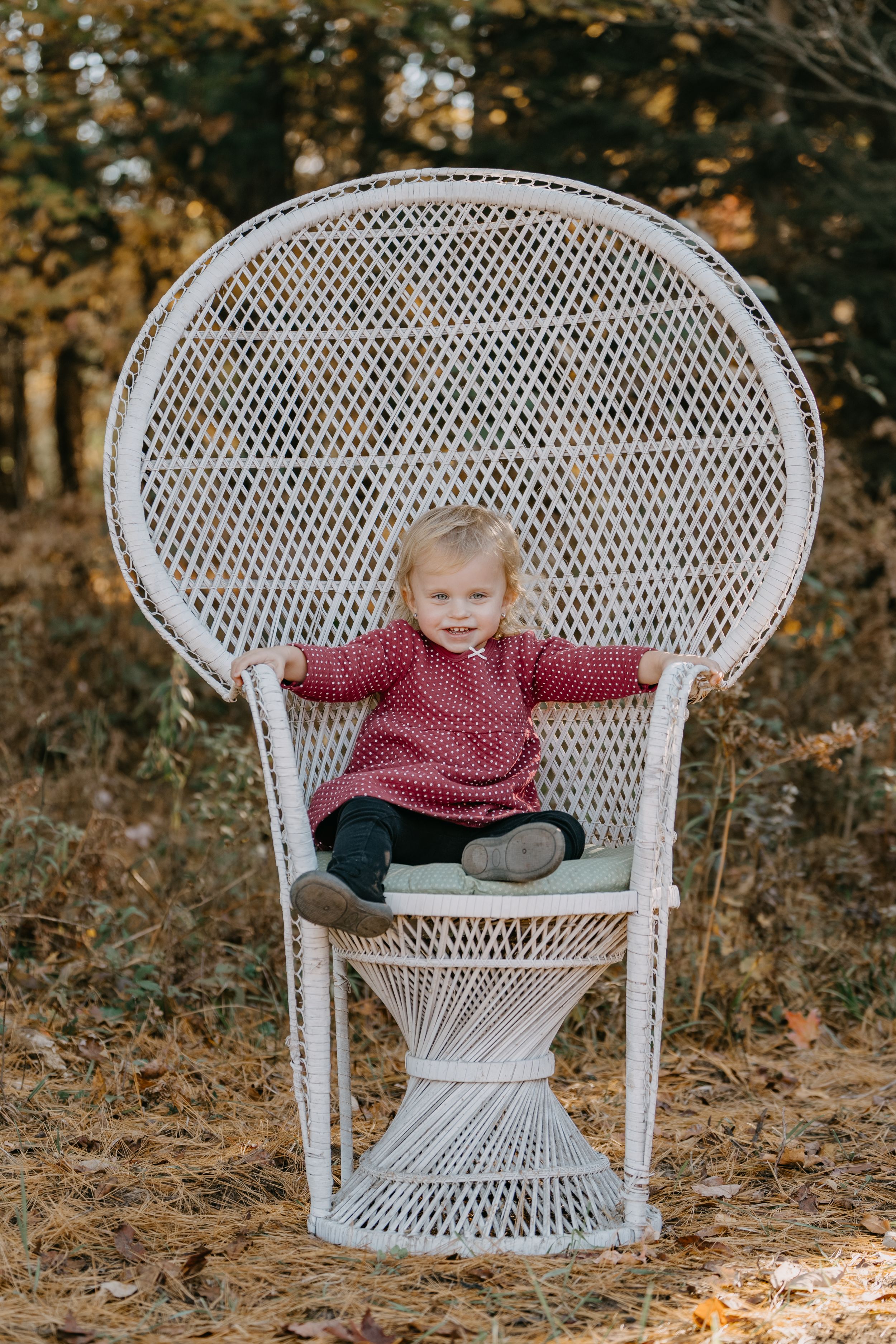 A small child wearing a red polka dot dress sits in a white wicker peacock chair outdoors in autumn.