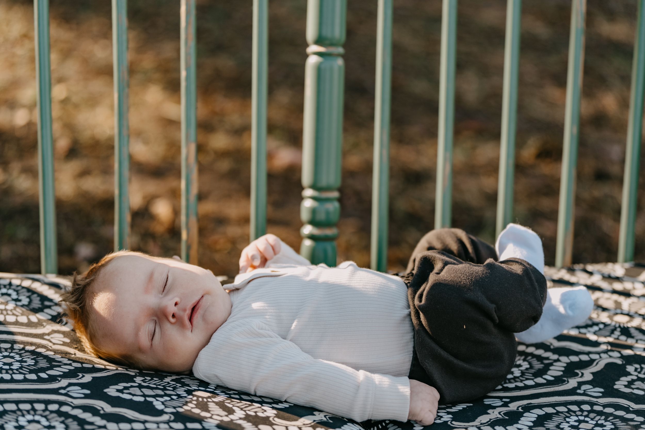 Baby sleeps peacefully outdoors on a patterned blanket next to blue metal railings on a sunny afternoon.