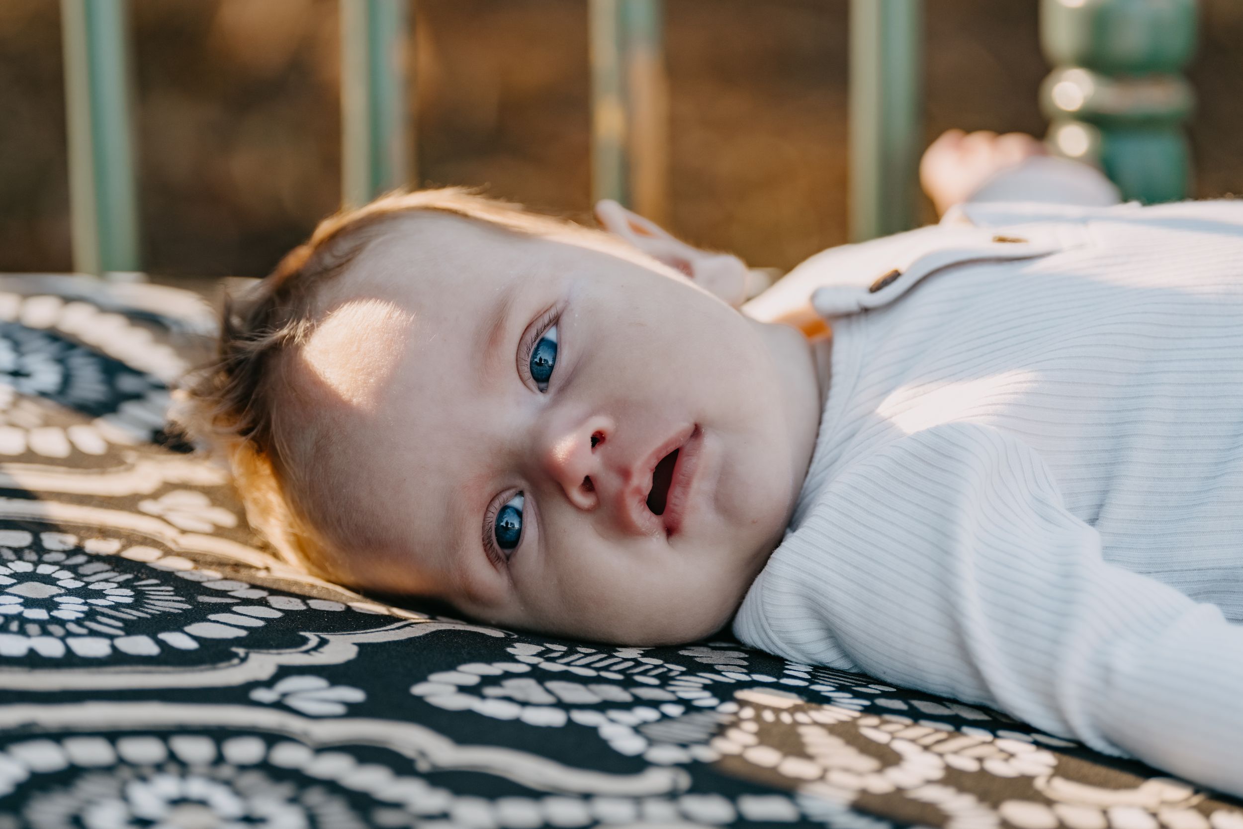 A baby in a white onesie lies on a patterned blanket looking up with bright blue eyes.