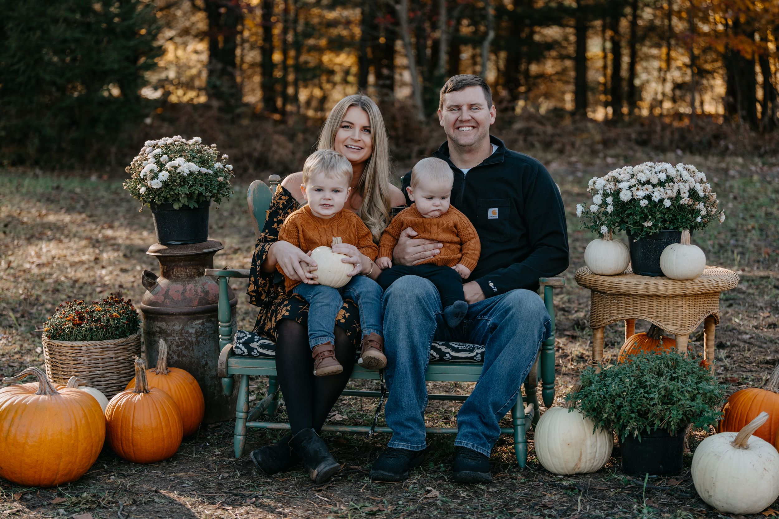 Family sits on rustic bench surrounded by fall decorations including white and orange pumpkins and mums in autumn setting.