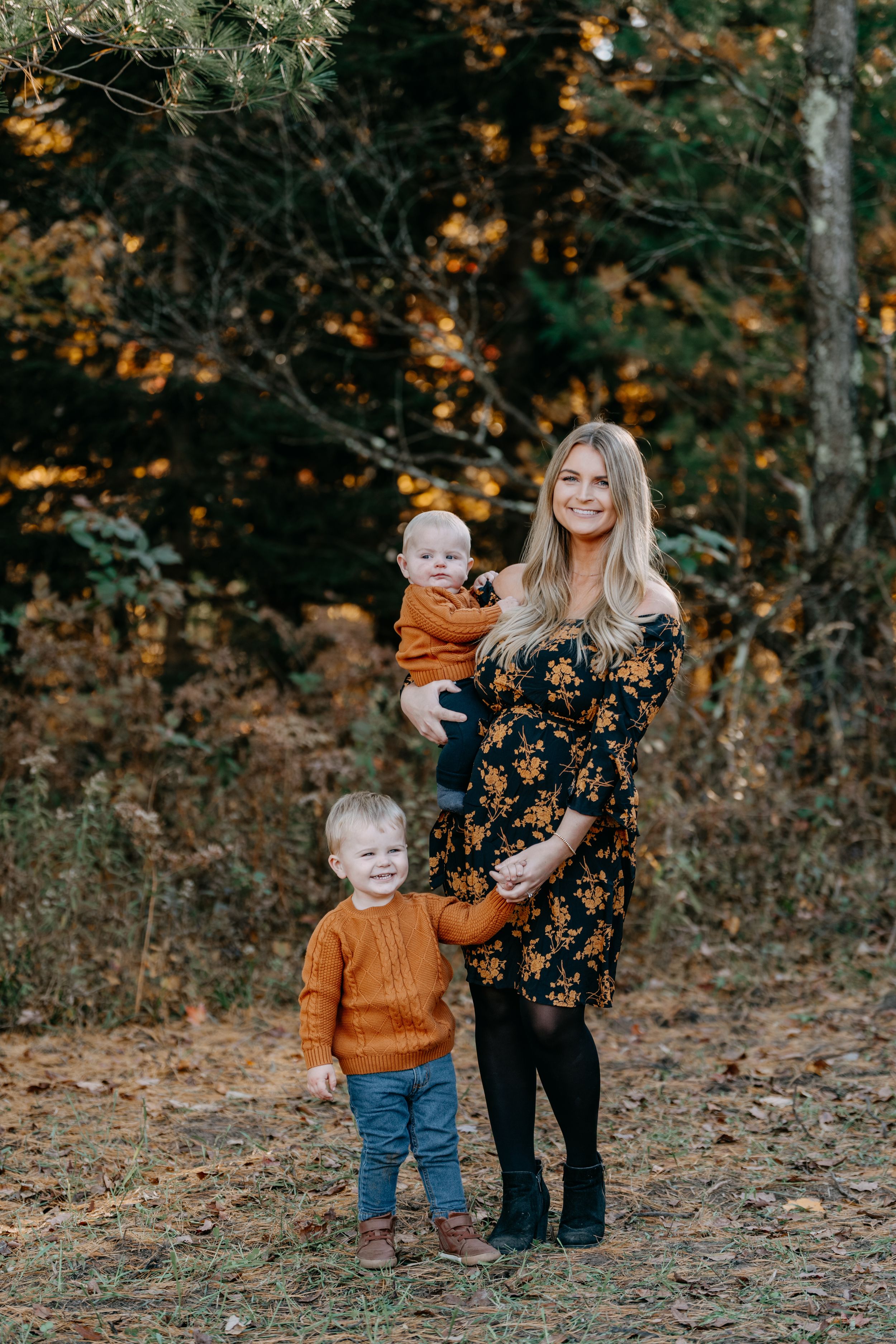 A family wearing autumn-colored clothing smiles and poses together in a wooded setting with fall foliage in the background.
