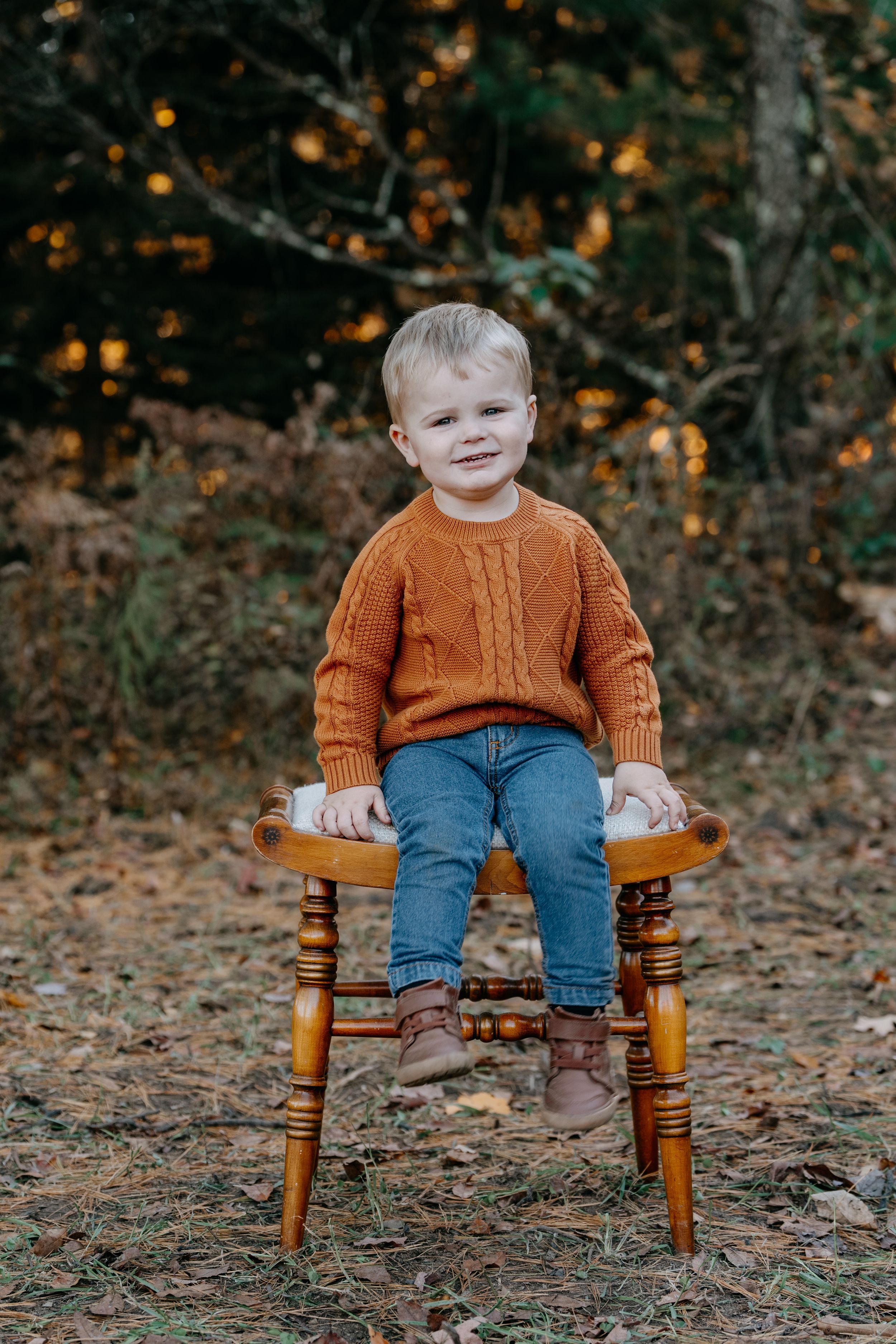 A small child in a rust-colored sweater and jeans sits on a wooden stool outdoors during autumn.