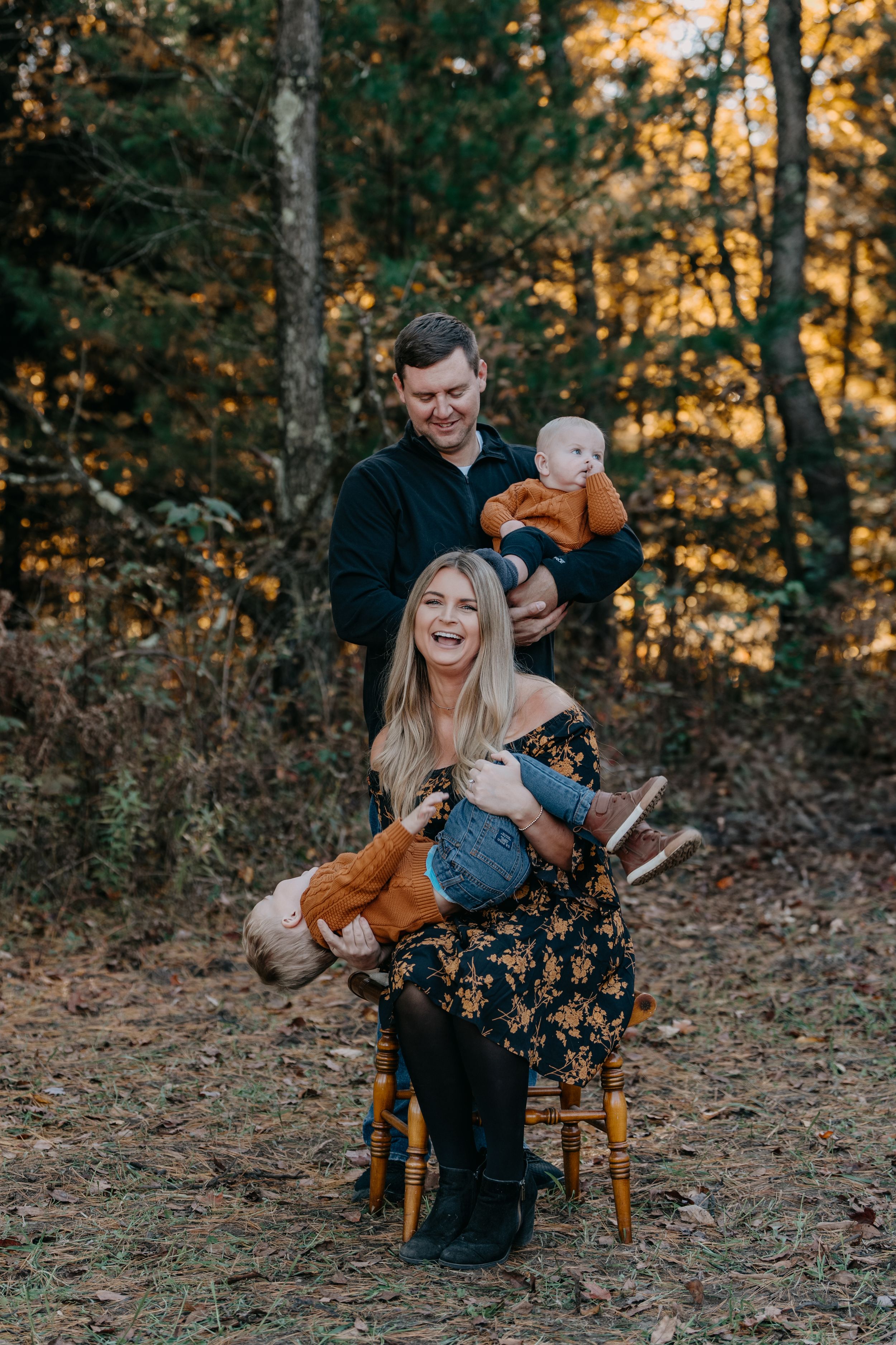 Family poses together in a wooded setting during autumn with golden sunlight filtering through the trees.