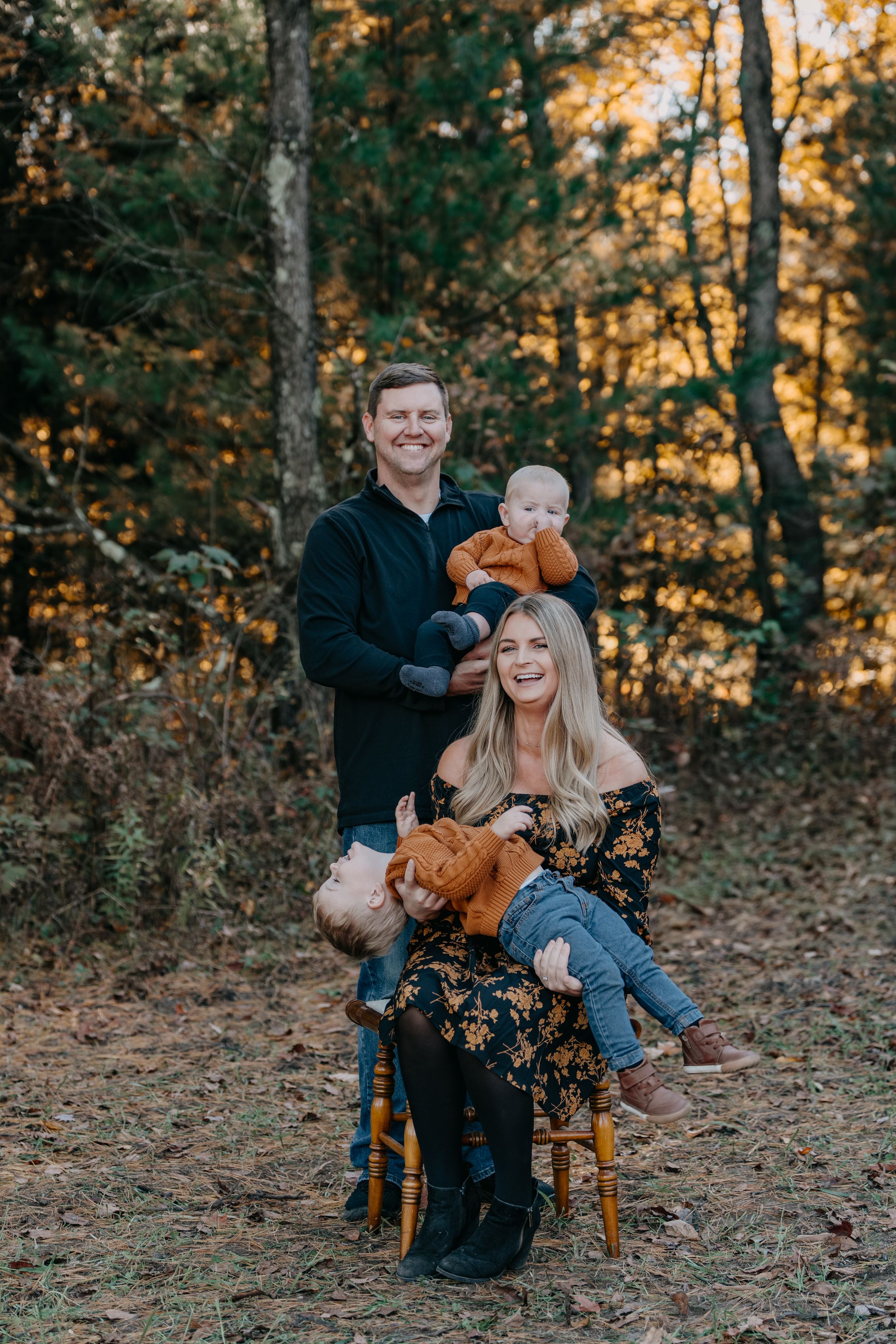 A playful autumn photoshoot in the woods shows two people laughing and sitting on chairs surrounded by fall foliage.