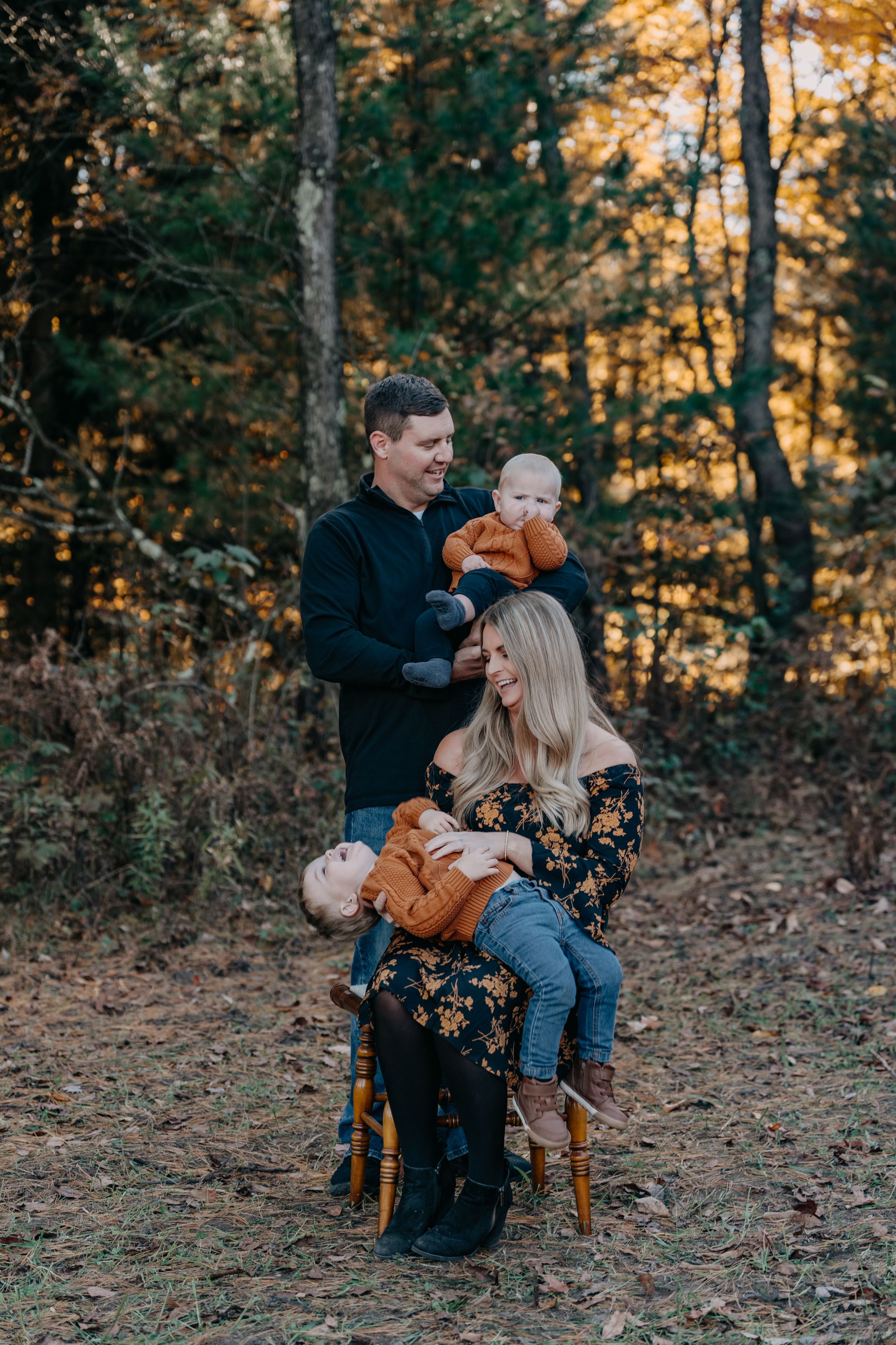 A family shares a tender moment in autumn woods while sitting on a wooden chair among fallen leaves at sunset.