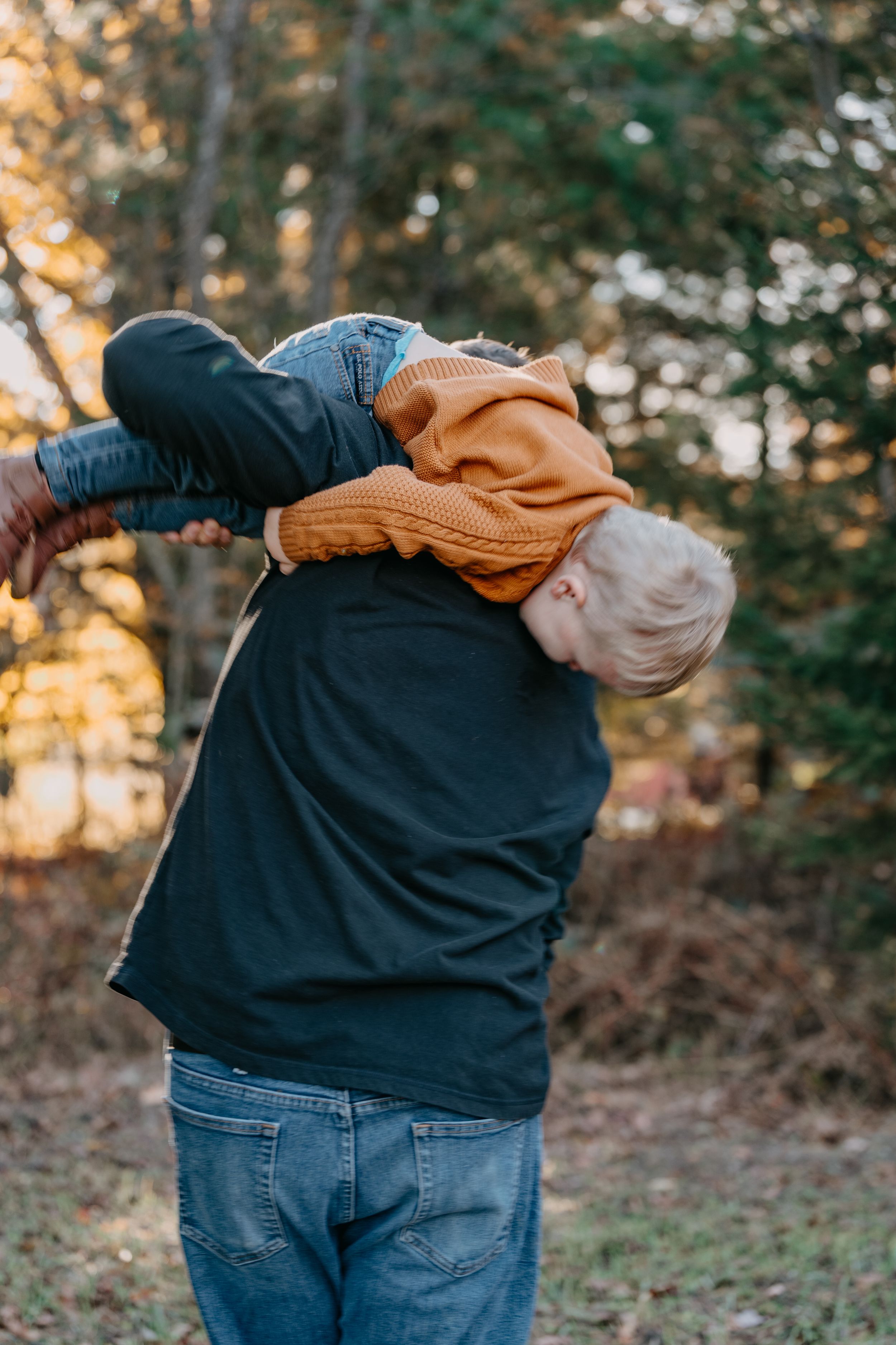 A parent wearing jeans and a navy sweater holds a child in a brown coat outdoors during golden hour in autumn.